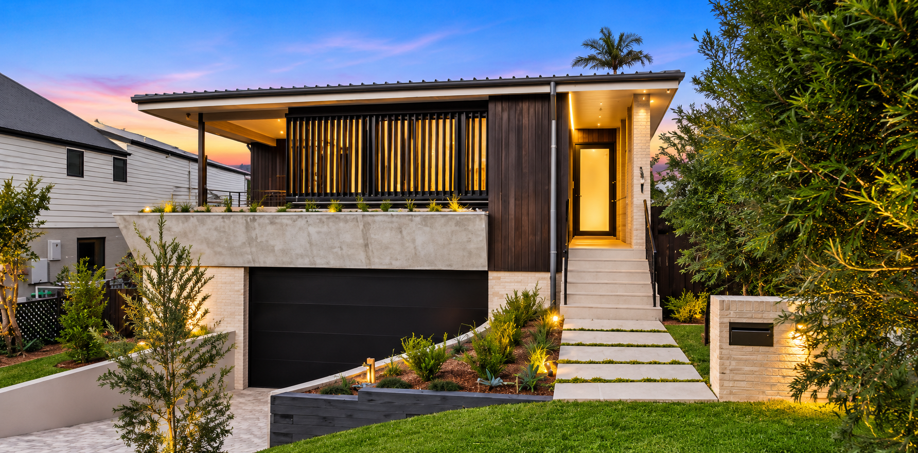 Modern house with exterior lighting at sunset, featuring a balcony with wooden slats, a staircase, landscaped garden, and a black garage door.