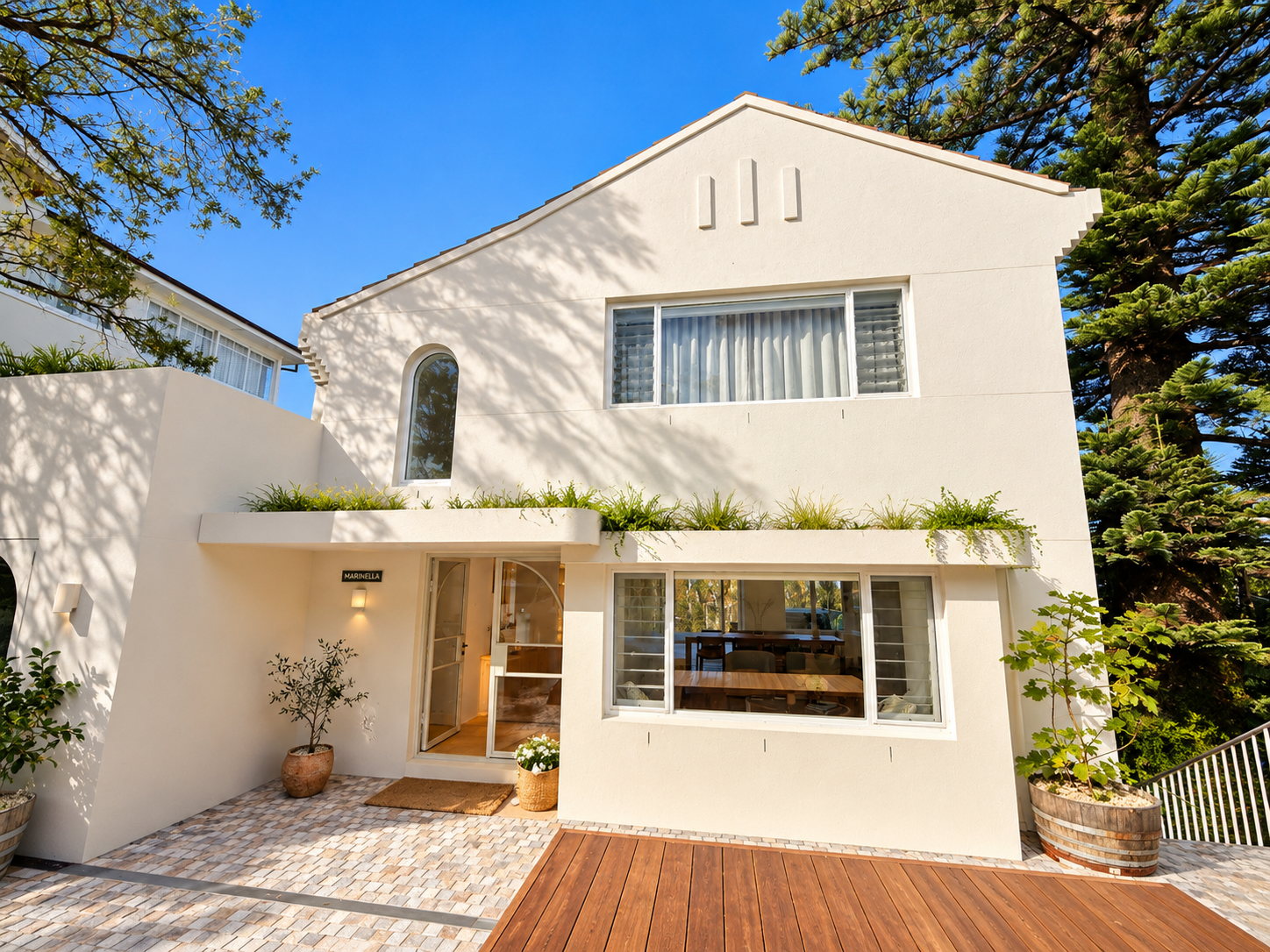 White multi-story house with large windows, surrounded by greenery and trees, under a clear blue sky.