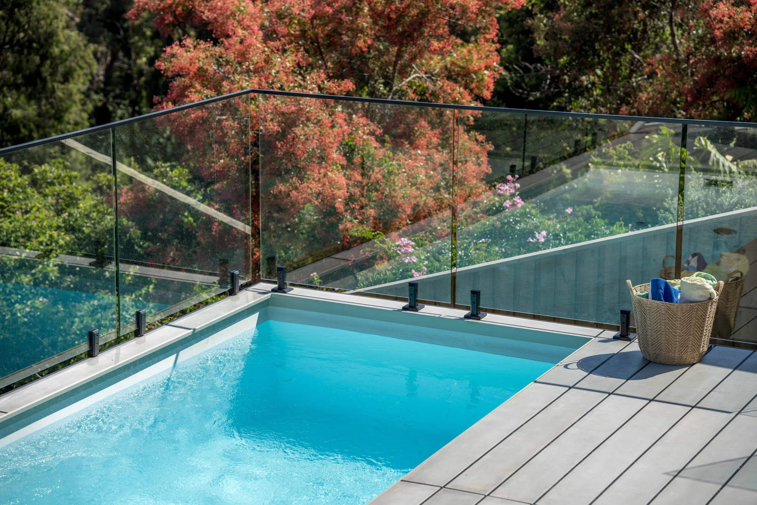 A swimming pool with clear blue water on a patio, surrounded by a glass railing and overlooking a lush garden with trees in autumn colors.
