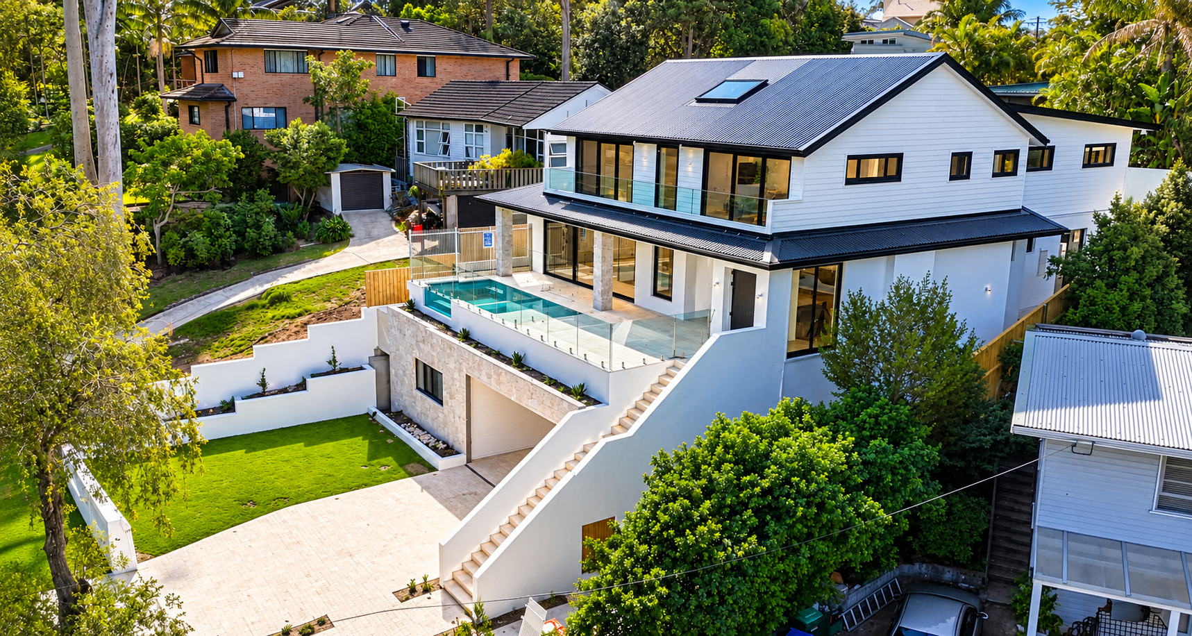 Modern multi-story house with a rooftop pool, glass fencing, and outdoor seating area, surrounded by lush greenery and neighboring homes.