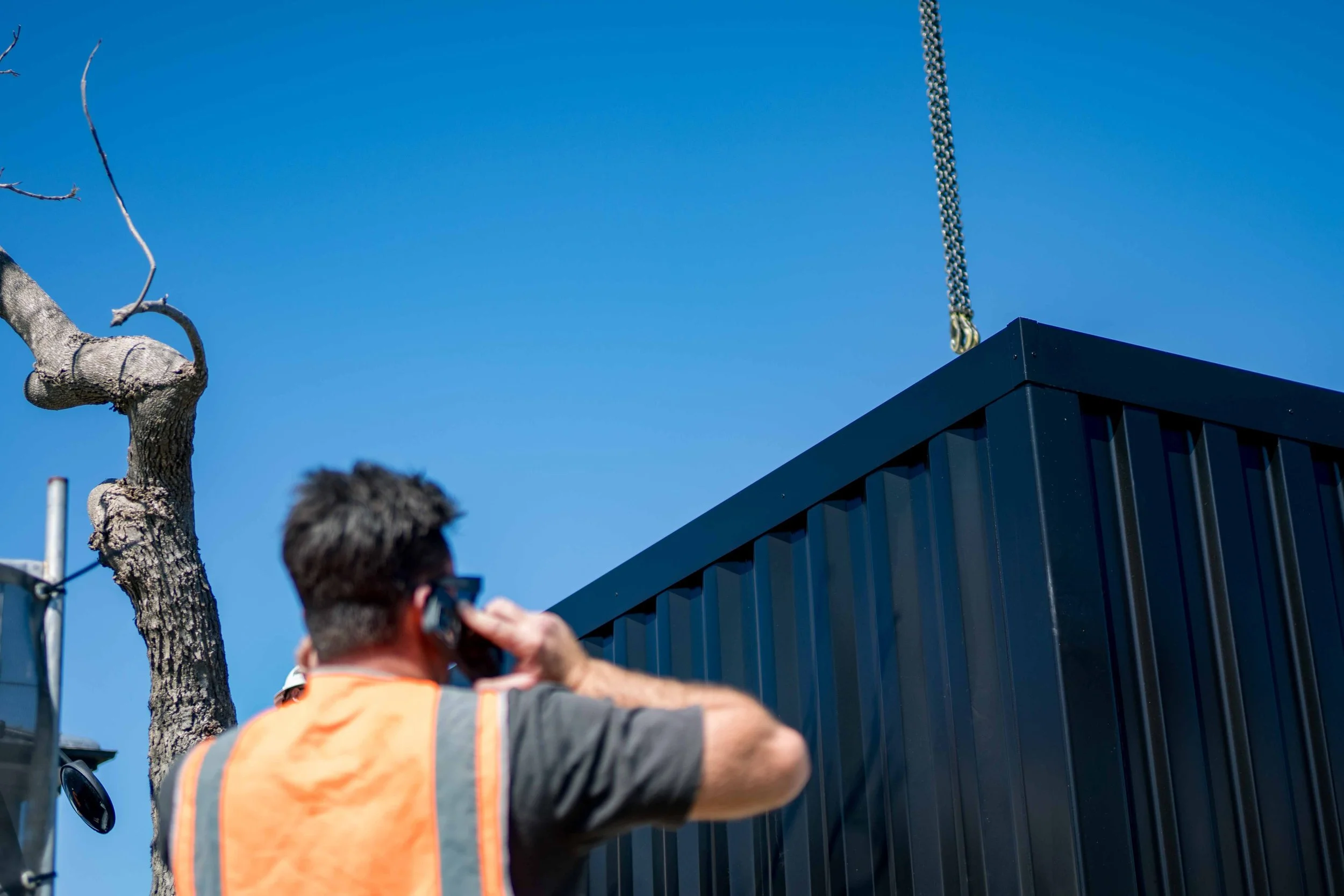 A person in an orange safety vest taking a photo of a black building with a chain hanging from its roof, a tree with bare branches nearby, under a clear blue sky.