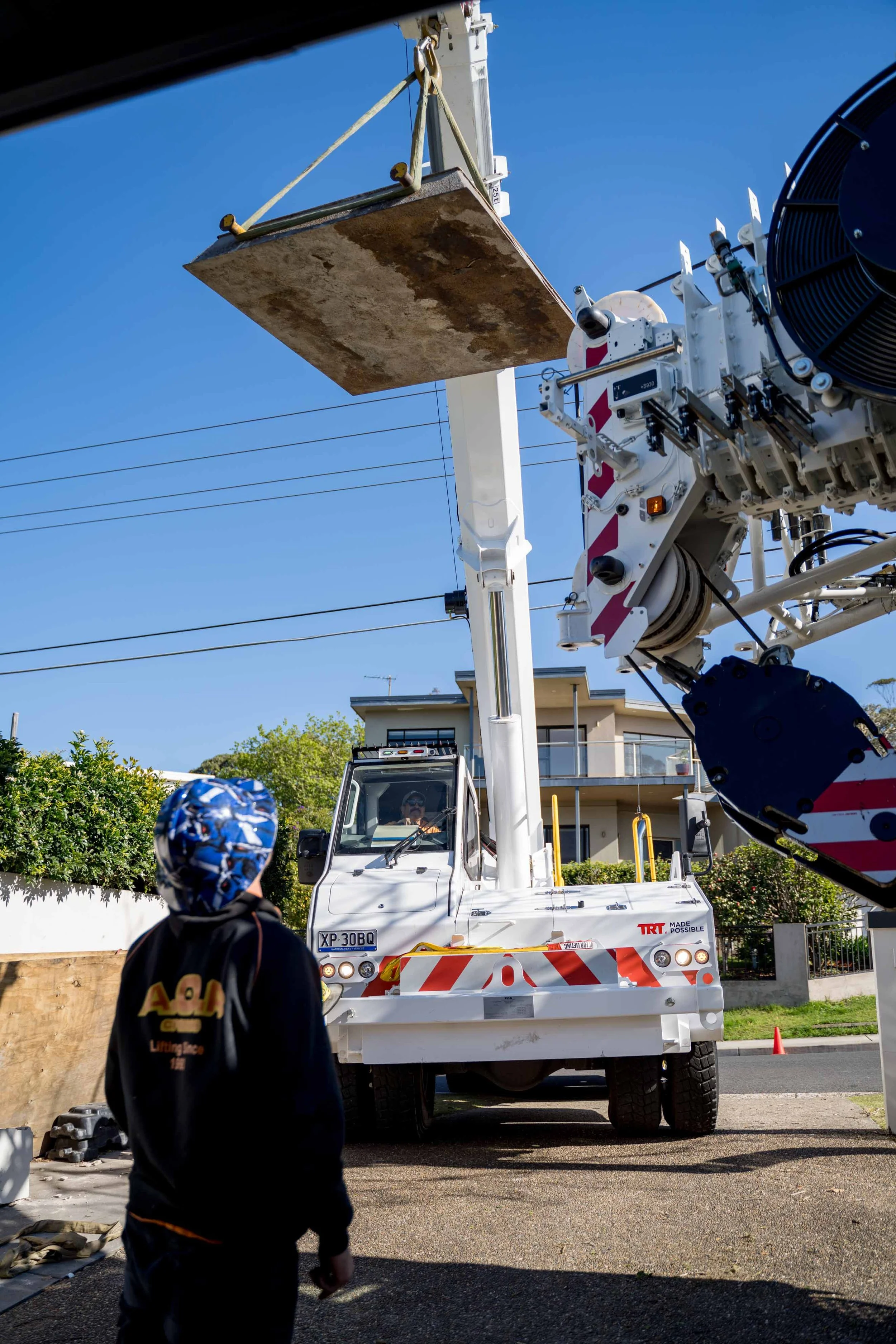 Person in black jacket and blue patterned cap watching a large white truck with a crane lifting a rectangular piece, under a clear blue sky.