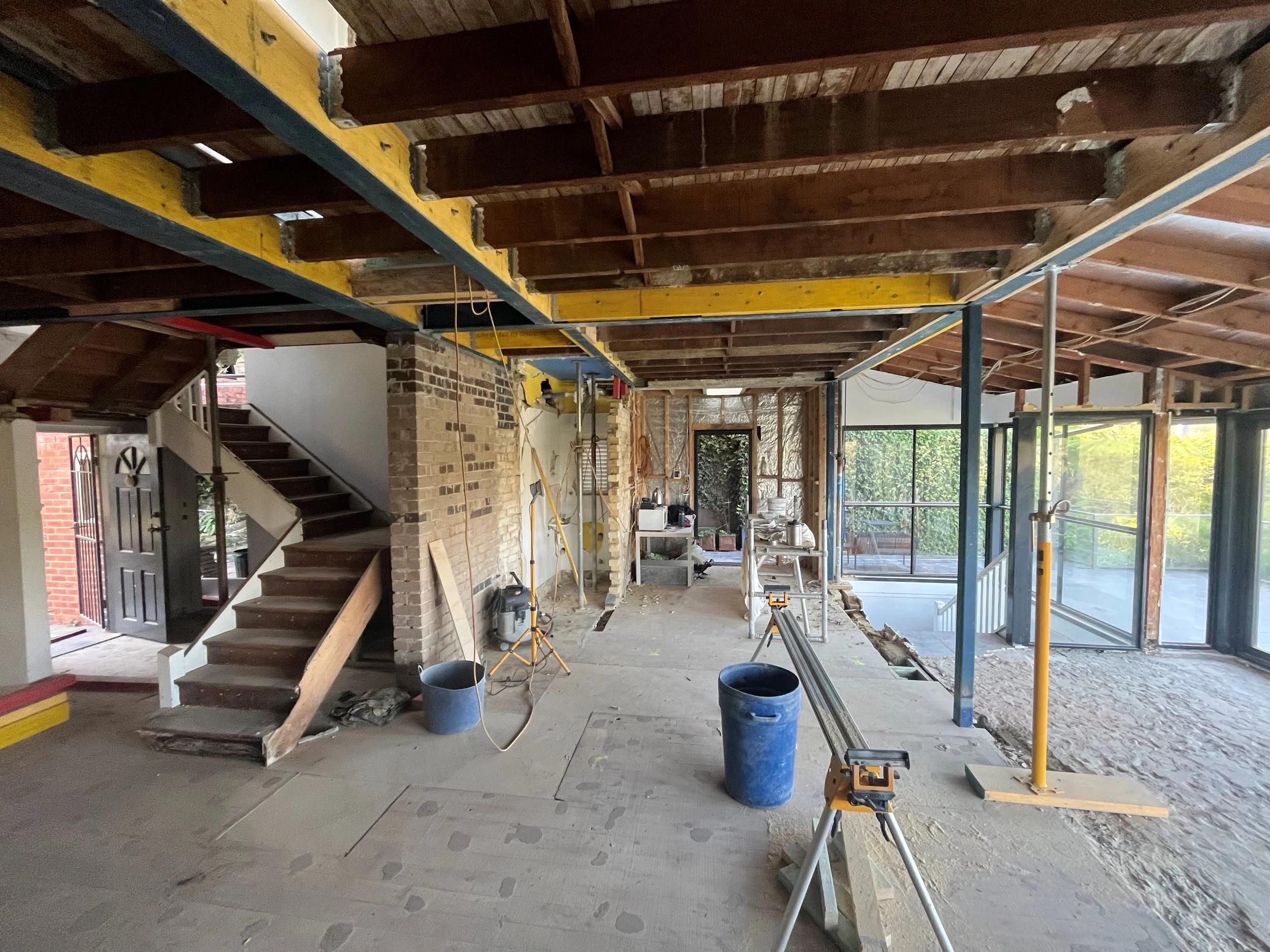 The interior of a house under renovation showing exposed wooden floors, a staircase, construction tools, scaffolding, and building materials.