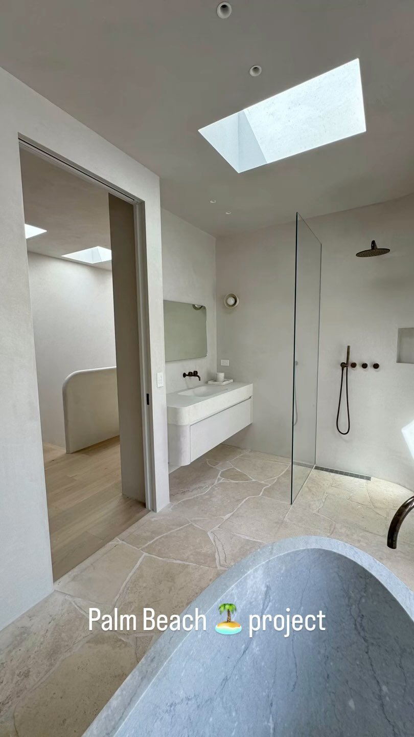 Modern bathroom with skylight, walk-in shower with black fixtures, floating white vanity, large stone bathtub, and neutral color palette.