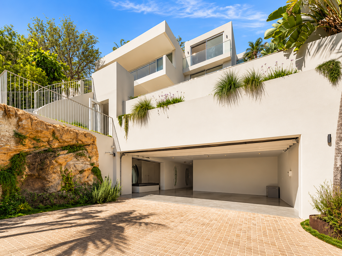 Modern white multi-level house with a garage, balcony, and greenery on a sunny day with a blue sky.