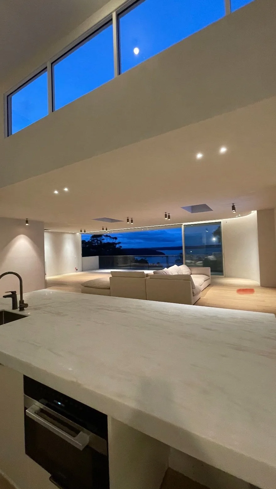 Modern living room with large windows showing a night view, white walls, a white sofa, and wooden flooring.