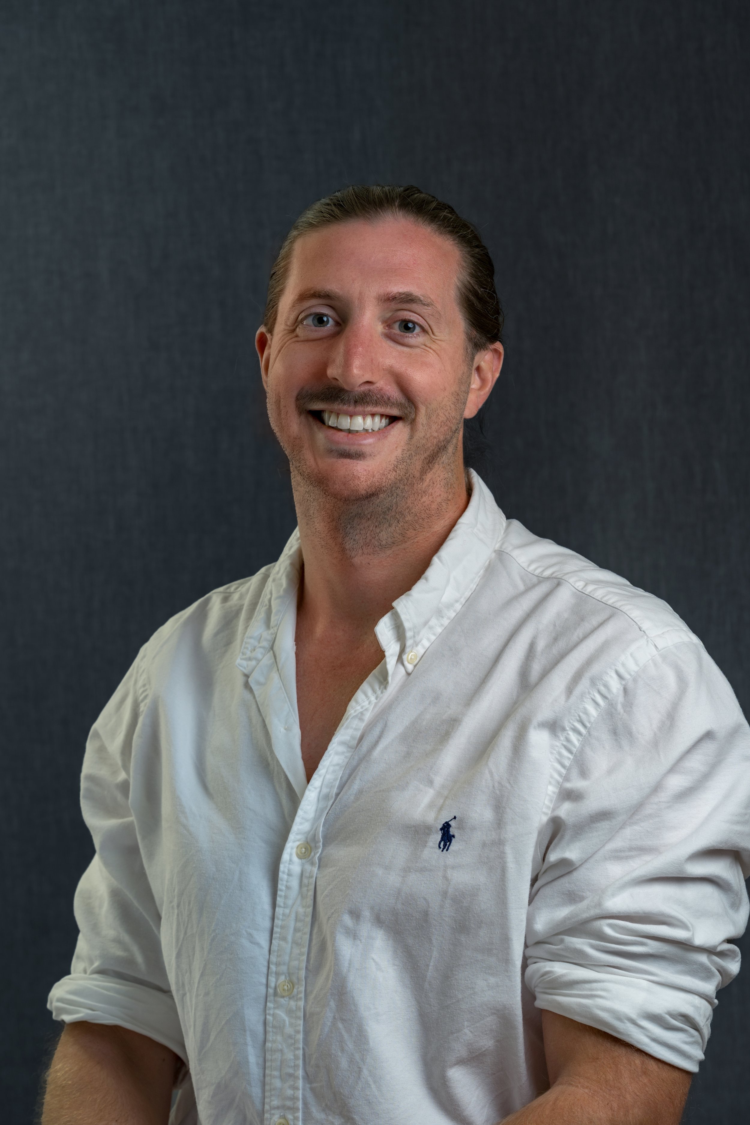 A smiling man with brown hair and a mustache wearing a white button-down shirt with a small blue logo on the left chest, posing against a dark grey background.