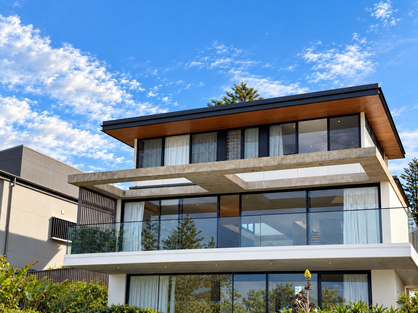 Modern multi-story house with large glass windows and balcony, under a blue sky with clouds.