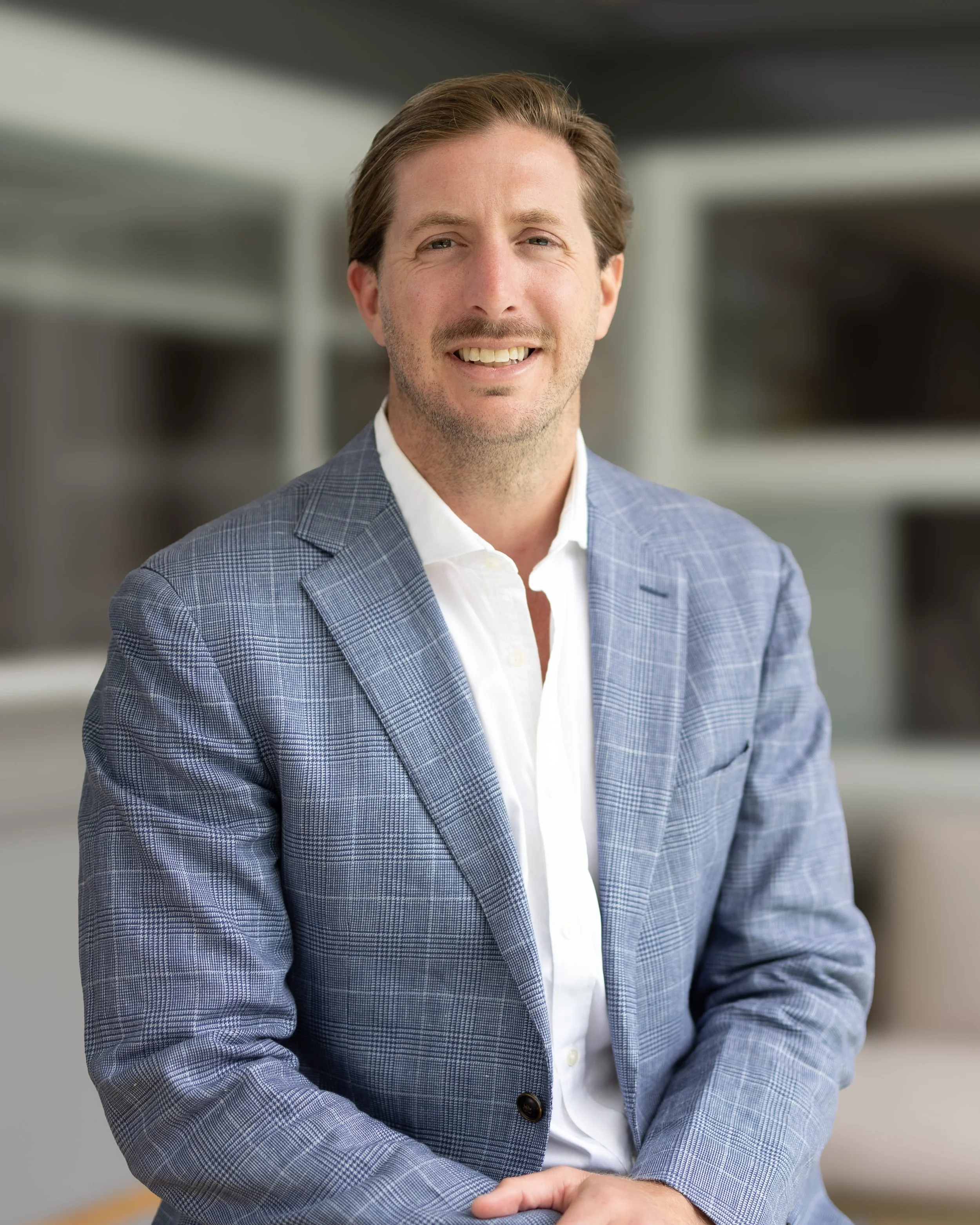 A smiling man in a blue checkered blazer and white shirt sitting in an office setting.