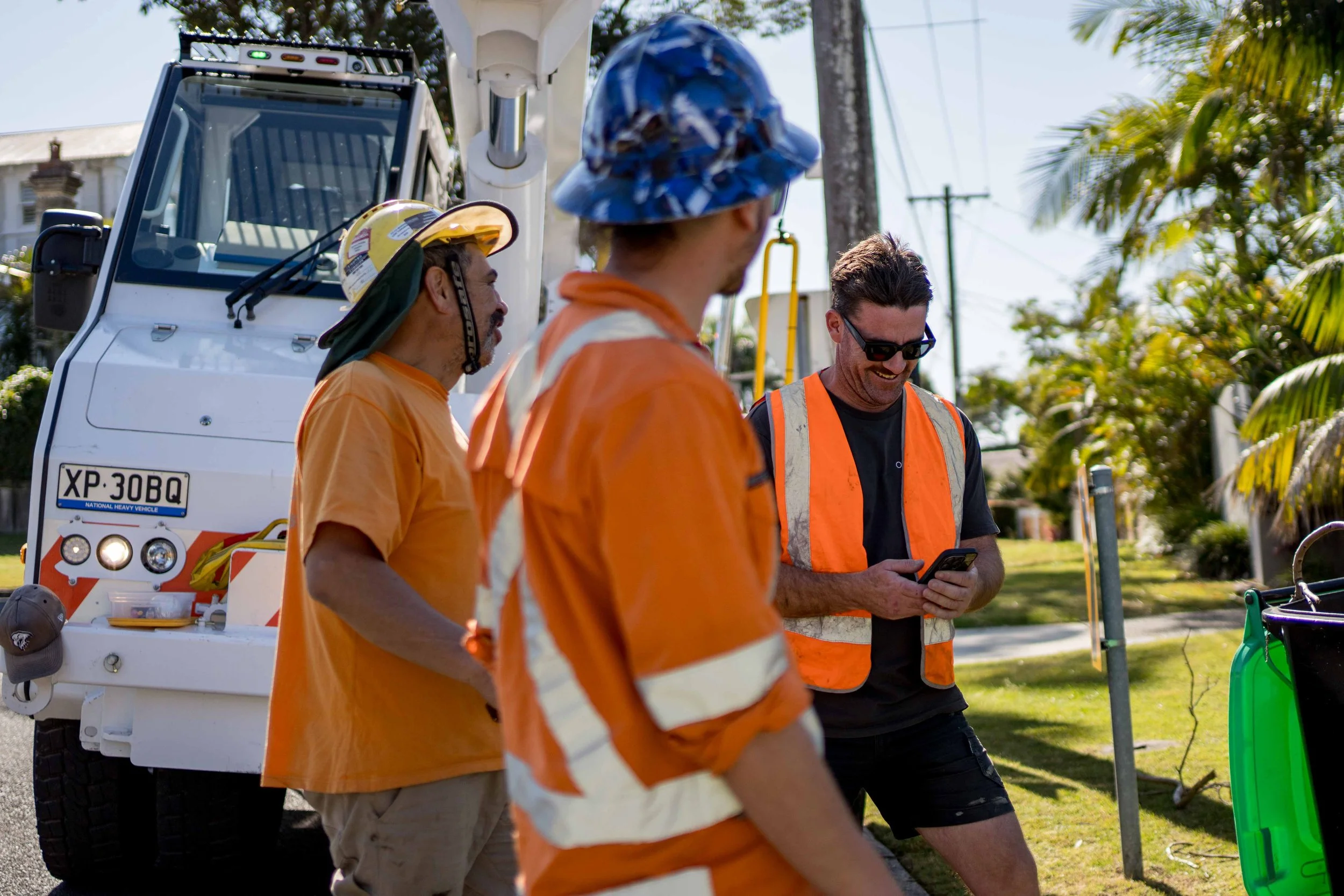 Group of utility workers in orange safety vests and hats standing outdoors near a utility truck, one man is using a handheld device.
