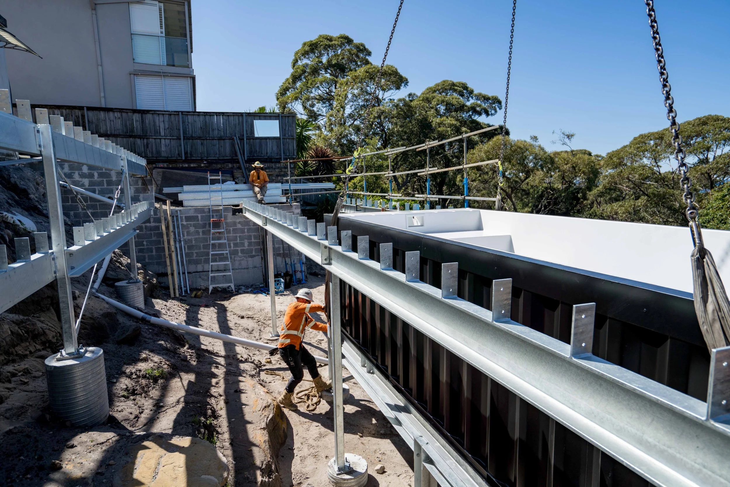 Construction workers installing structural steel beams on a building site, with trees and a building in the background.