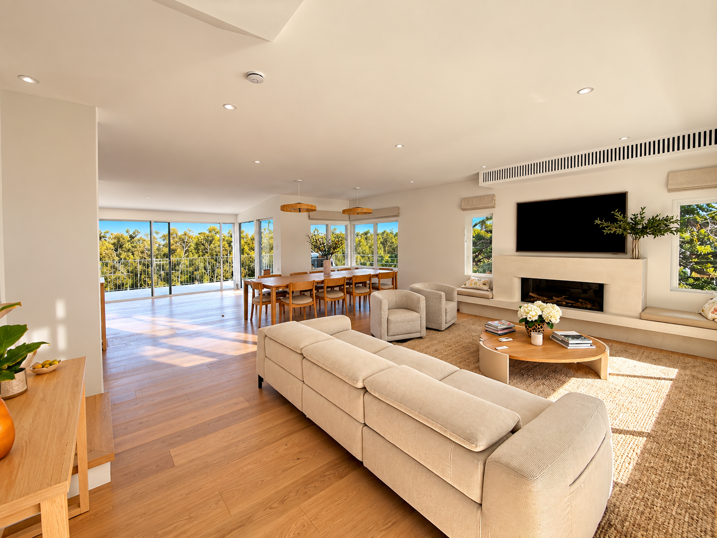Bright, modern open-concept living and dining room with large windows, hardwood floors, beige sofa, armchairs, a coffee table with flowers, and a mounted flat-screen TV above a fireplace.