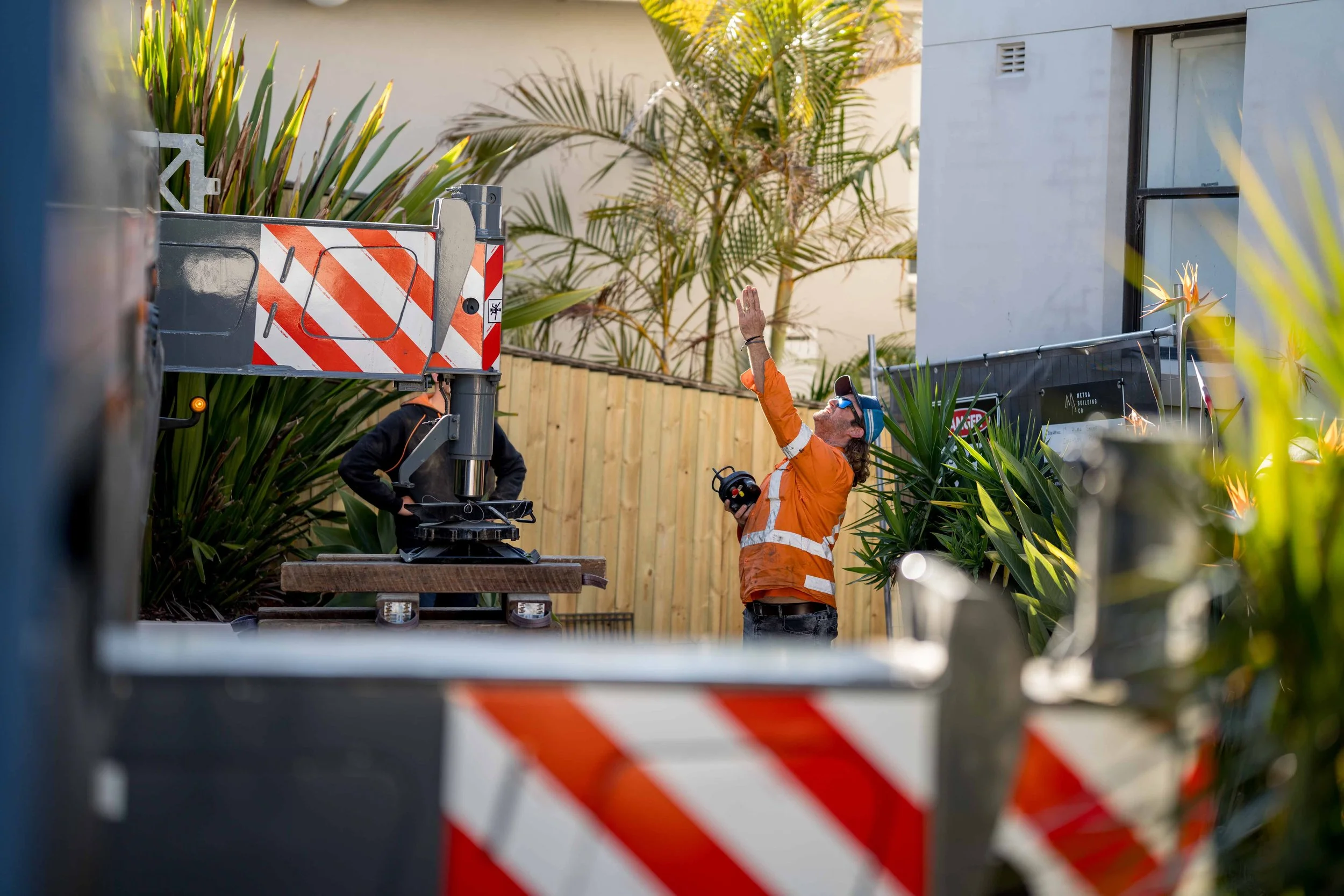 A construction worker in an orange safety vest and cap operating a machinery with a hand raised, surrounded by plants and fencing at a construction site.