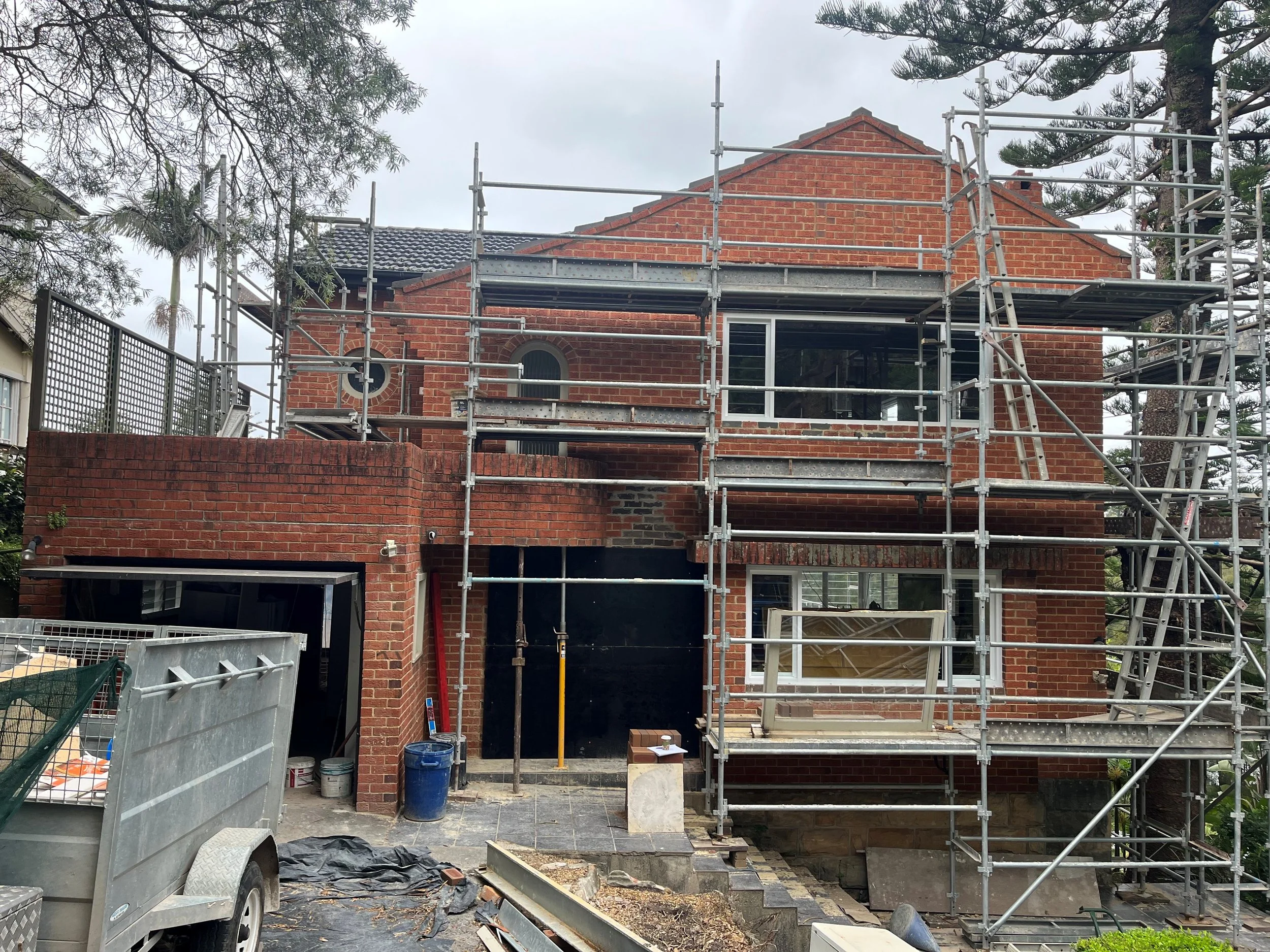 A brick house under construction with scaffolding surrounding it, construction materials and tools in the yard, and trees nearby.