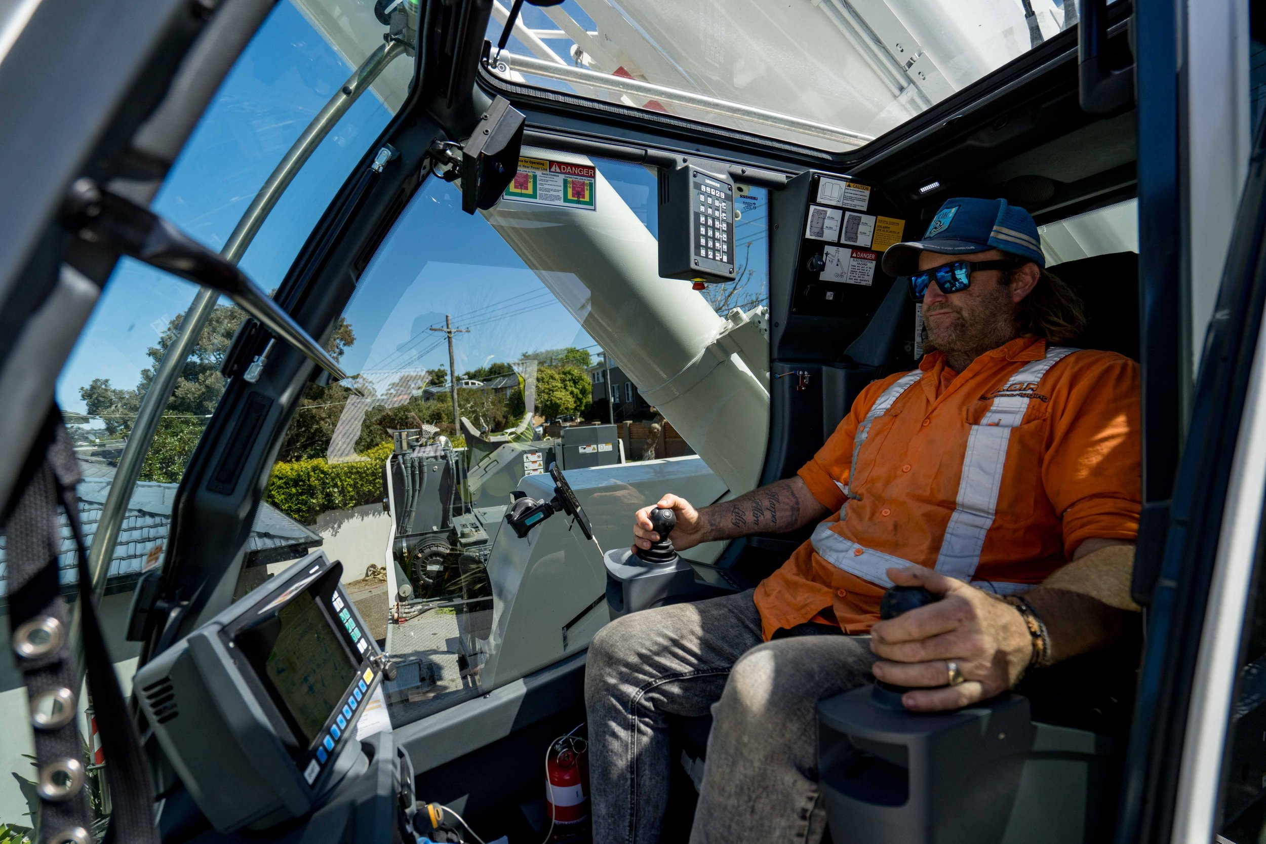 A man wearing sunglasses, a cap, and an orange work shirt is sitting inside a large construction machine, likely an excavator or crane. He appears to be operating the machine, holding a joystick with one hand and resting the other on his lap. The interior of the machine has control panels, screens, and warning labels, with outdoor scenery visible through the windows.