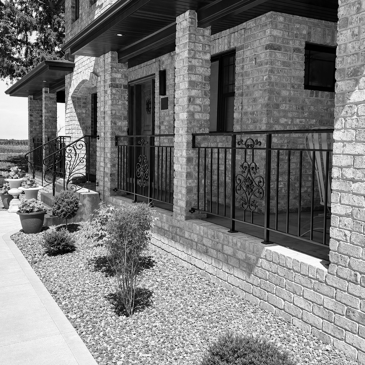 Black and white photo of a residential front porch with brick columns, wrought iron railings, and potted plants in a landscaped yard.