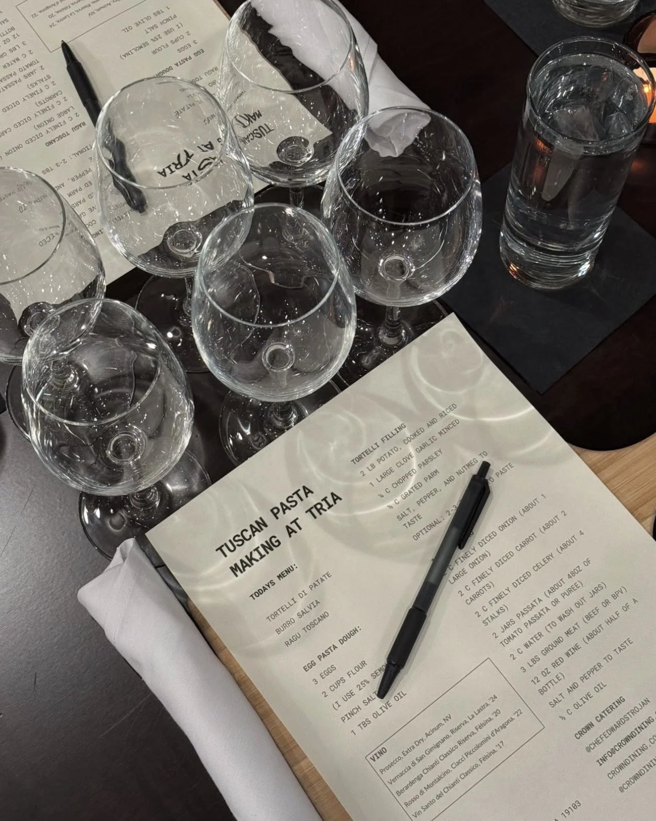 Multiple empty wine glasses and a glass of water on a table with a wine tasting menu and a pen nearby.