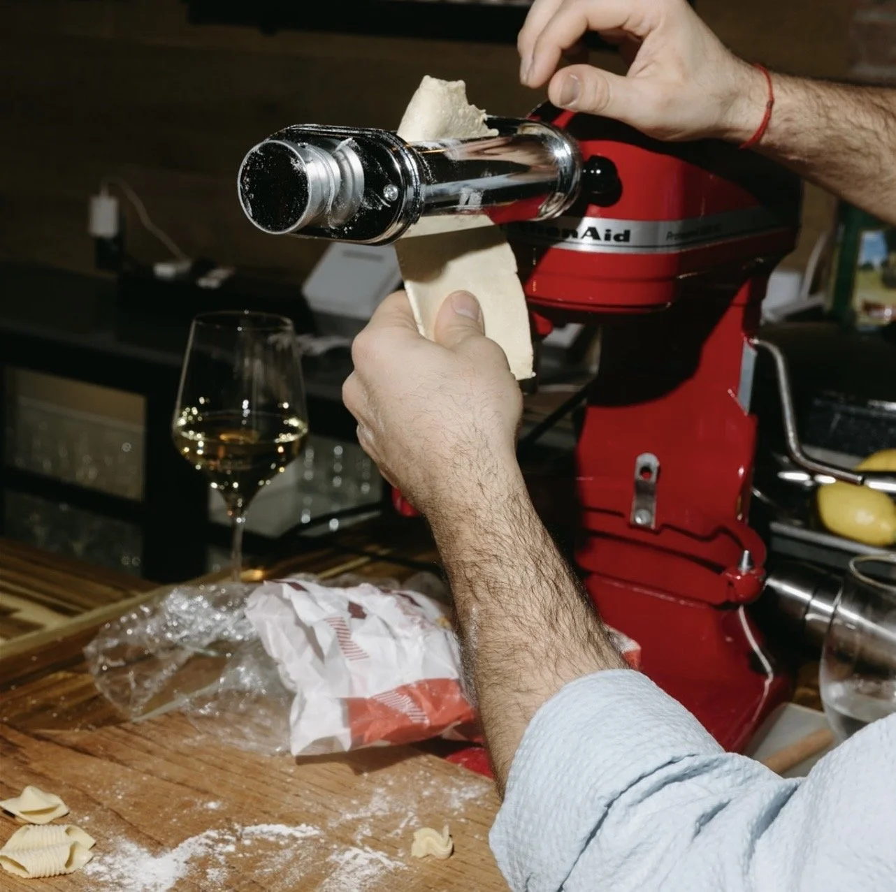 Person preparing freshly made pasta using a pasta machine, with a glass of white wine on the table.