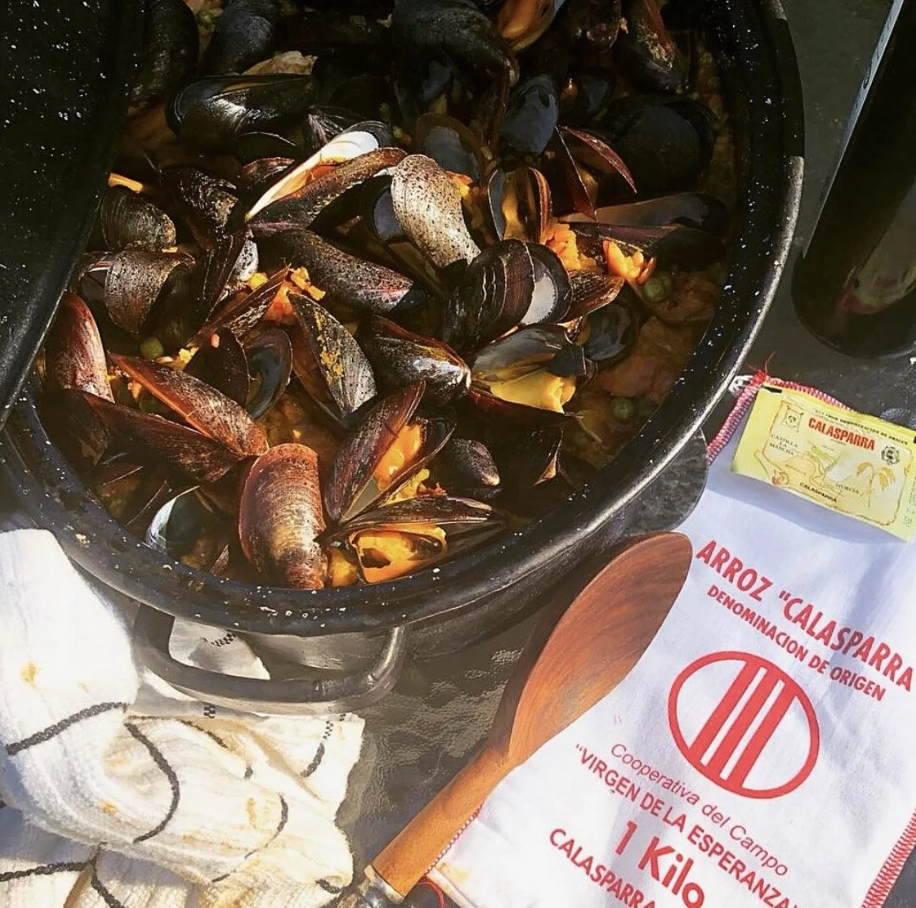 A black dish filled with cooked mussels and yellow-orange sauce, surrounded by a paper with red text about Calasparra rice, a wooden spoon, a packet of rice, a white towel, and a dark container.