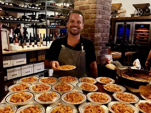 A smiling man in a black shirt and apron stands behind a table filled with plates of pasta, holding a plate of pasta in his right hand. Shelves with wine bottles and a wine fridge are visible in the background at a restaurant.