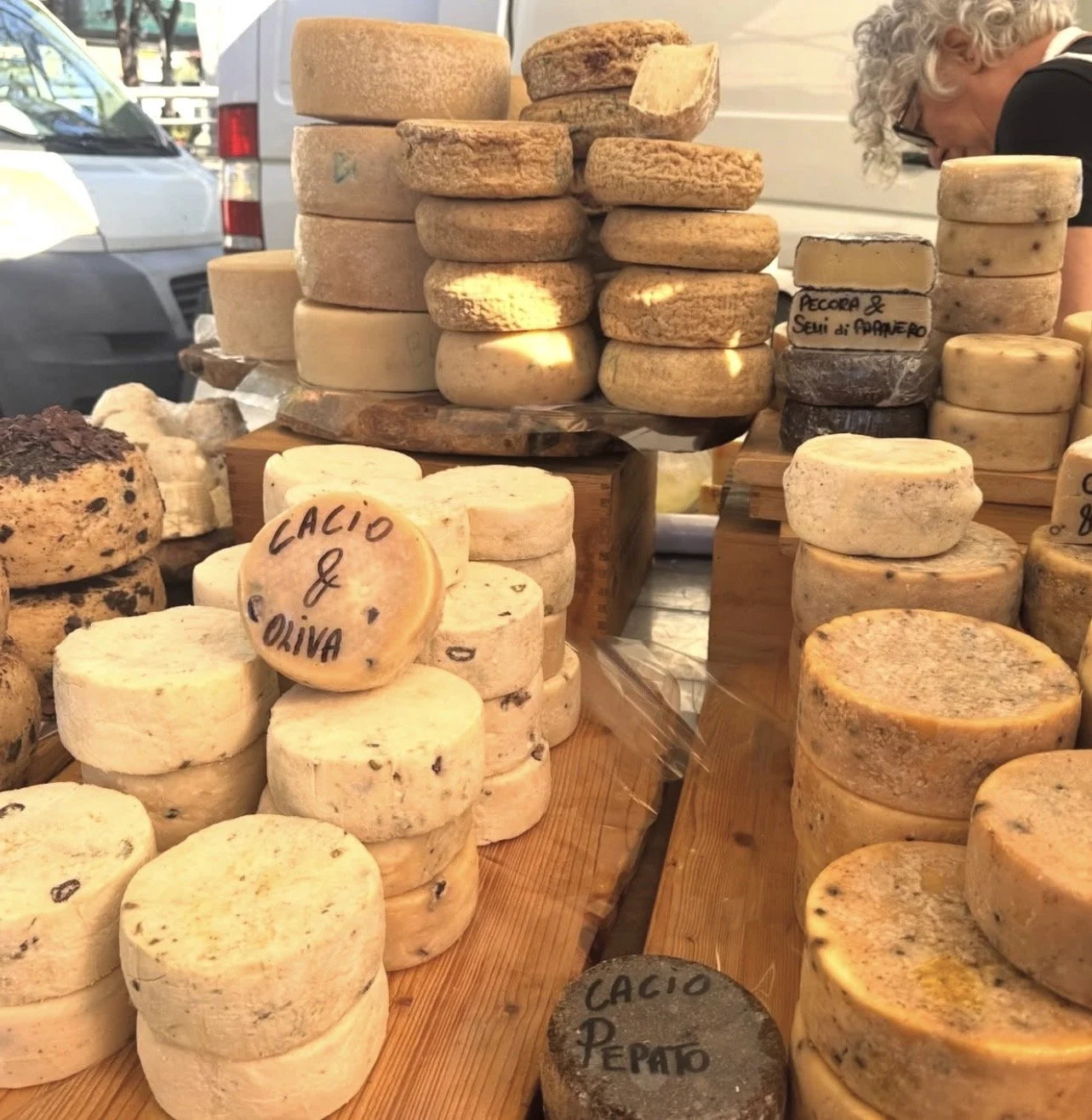 Various types of cheese stacked on wooden display tables at an outdoor market, with handwritten labels including 'Laccio & Oliva' and 'Pepe & Semi di Papavero'.