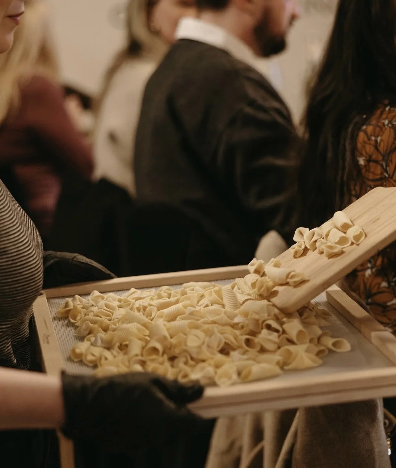 Person holding a tray of freshly made pasta shells, in a busy kitchen or pasta-making setting.