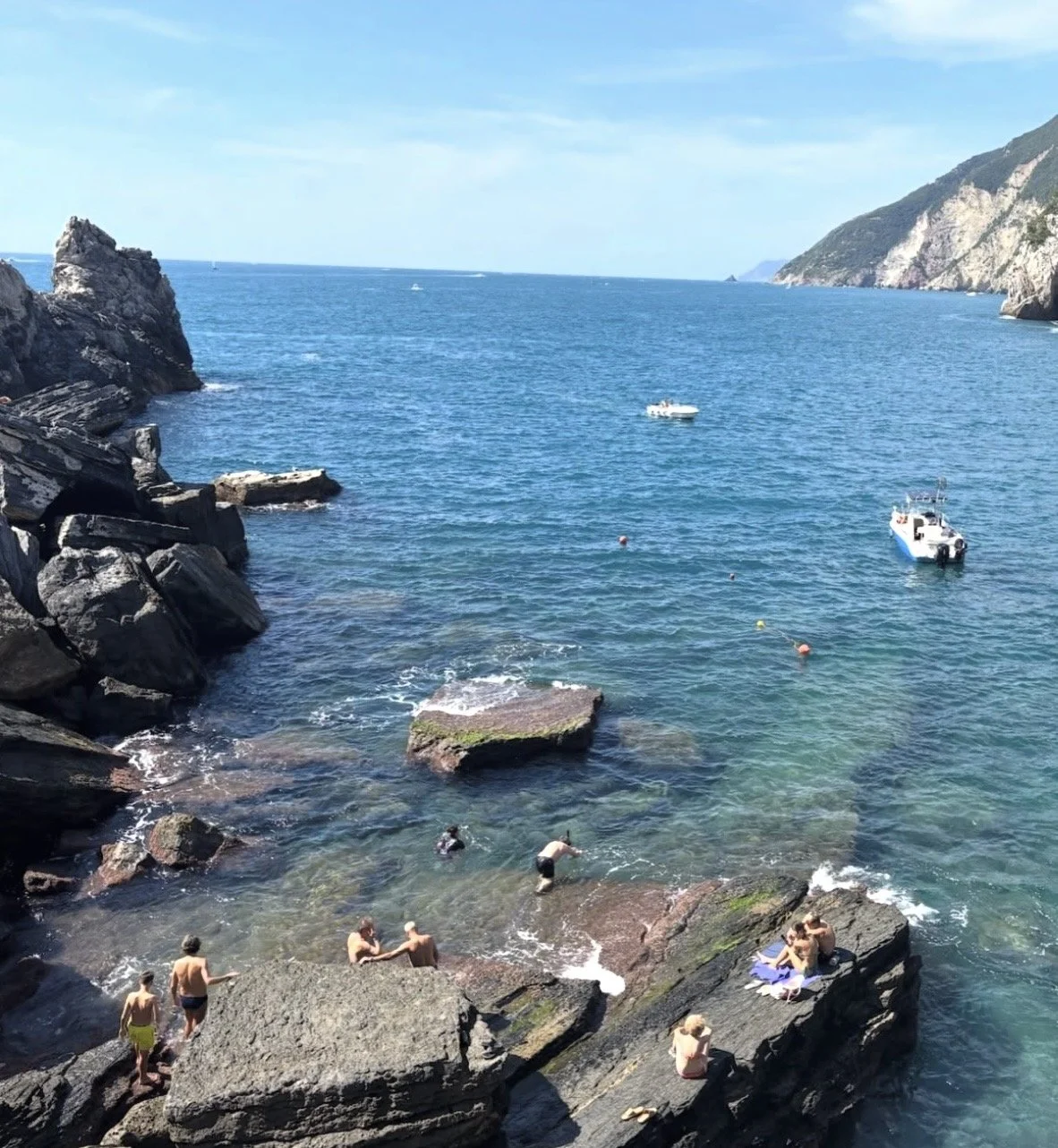 People swimming and relaxing on rocks by the ocean near boat and distant hills under clear blue sky.