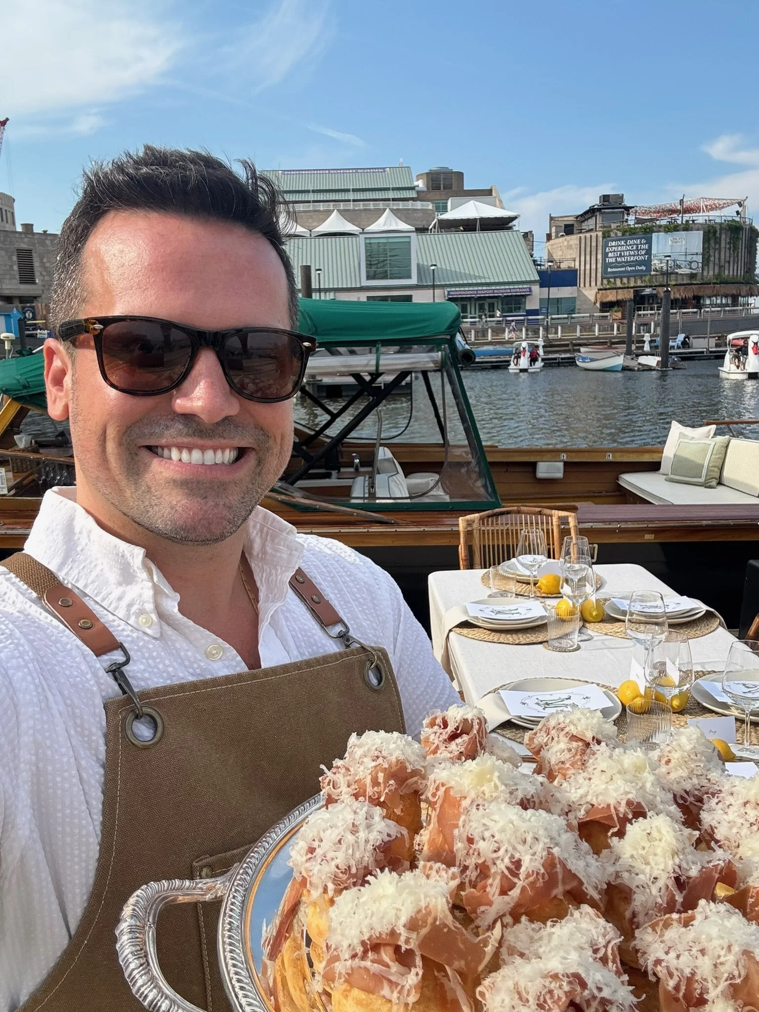 Smiling man wearing sunglasses, white shirt, and apron, sitting at an outdoor dining table by the water with a waterfront scene in the background, including boats, buildings, and blue sky with clouds.