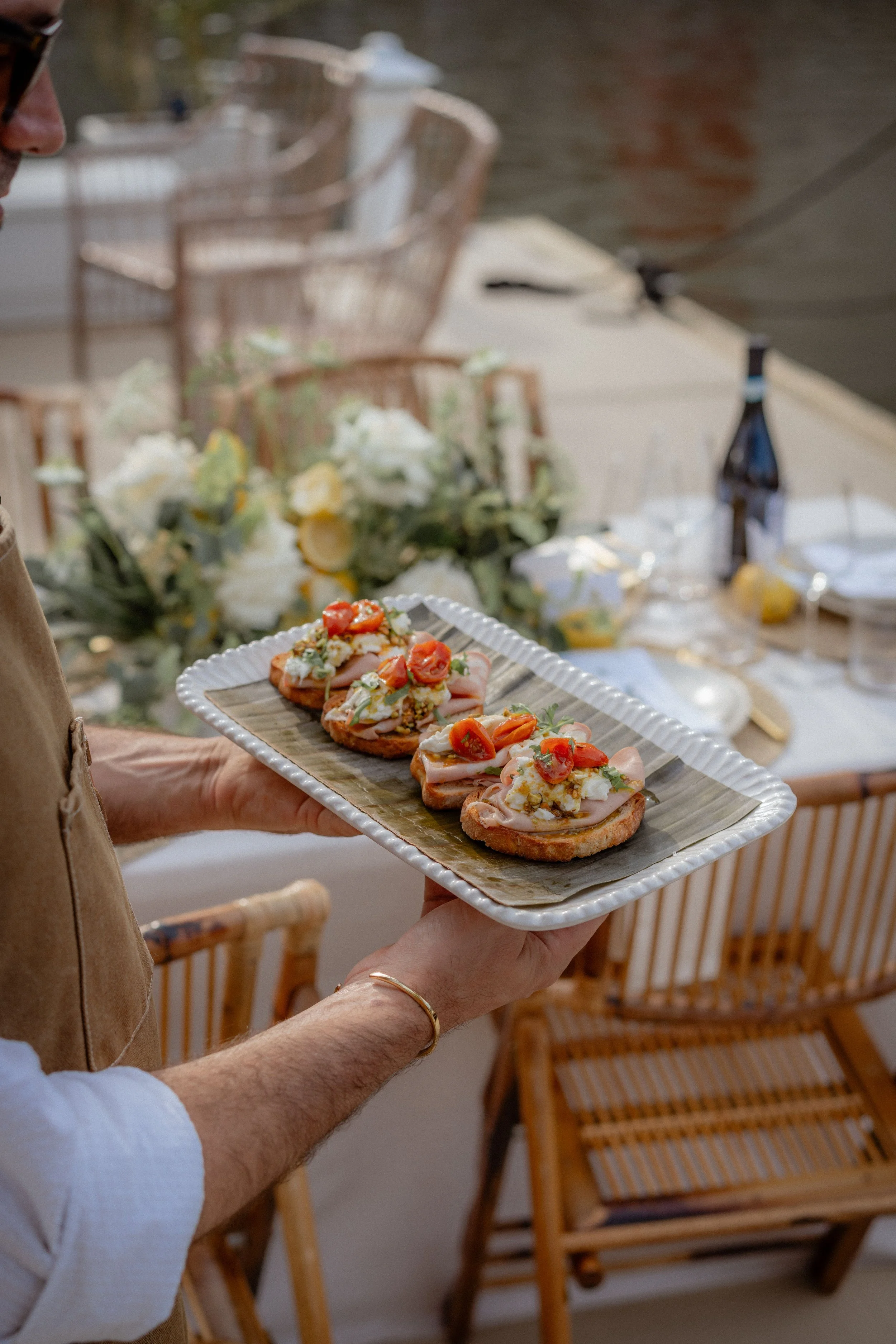 Person holding a rectangular tray with four open-faced sandwiches topped with cherry tomatoes, herbs, and cheese, at a dining table with a floral centerpiece and chairs.