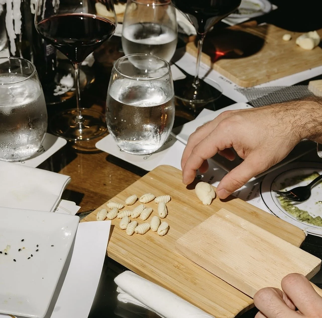 A person is placing a piece of gnocchi on a wooden cutting board with other gnocchi. There are wine glasses and water glasses on the table, along with plates of food, including a bowl with green sauce and a white dish with black pepper.