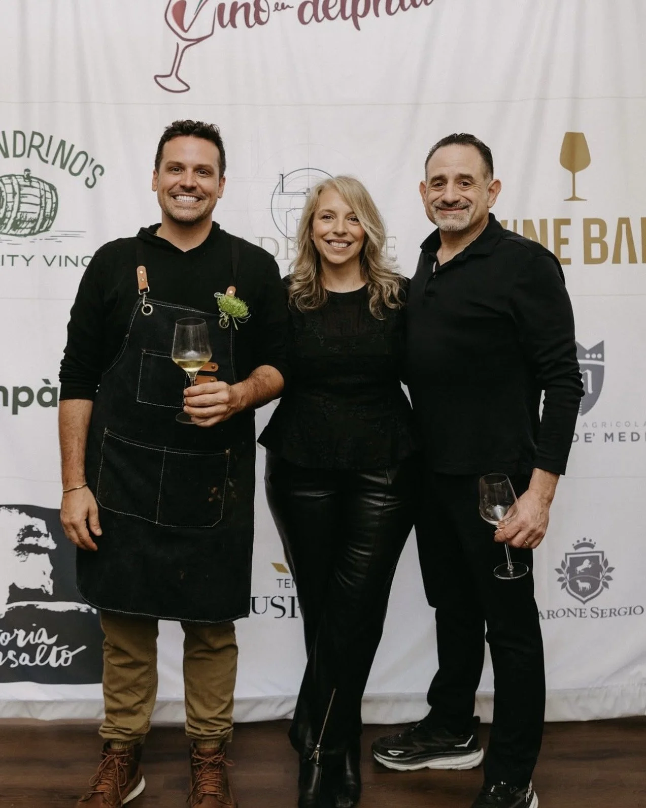 Three people standing together at an event with a white backdrop featuring wine-related logos and text. The person on the left wears a black apron and holds a glass of white wine. The woman in the middle wears a black top and leather pants. The person on the right wears a black shirt and pants, holding a wine glass. All three are smiling.