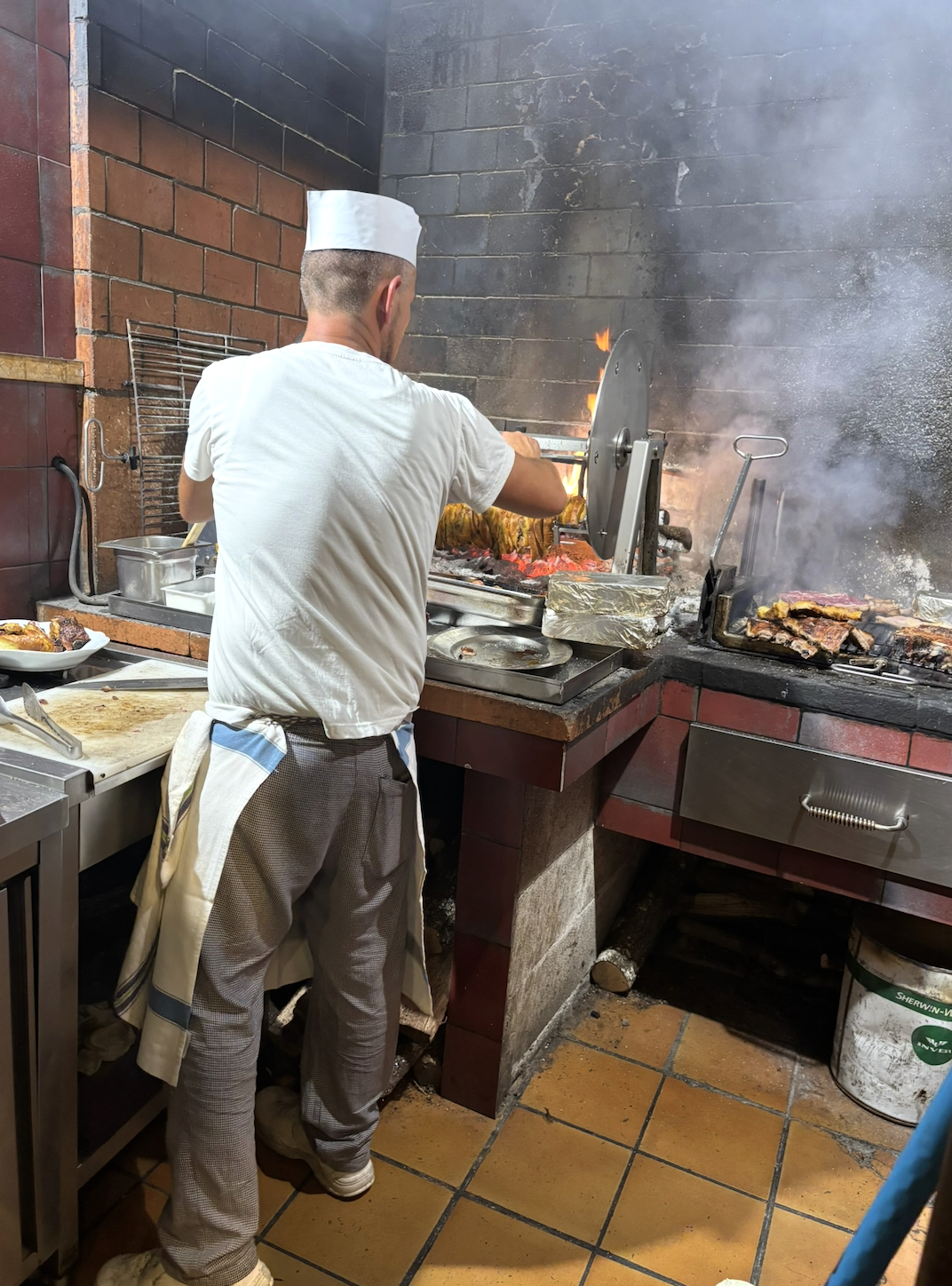 A chef in a white hat and apron cooks meat over an open flame grill at a restaurant kitchen.