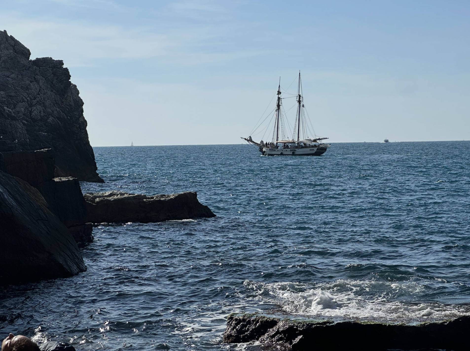 A sailboat with multiple masts sailing on blue ocean waters, with rocky coastline in the foreground and a clear sky above.