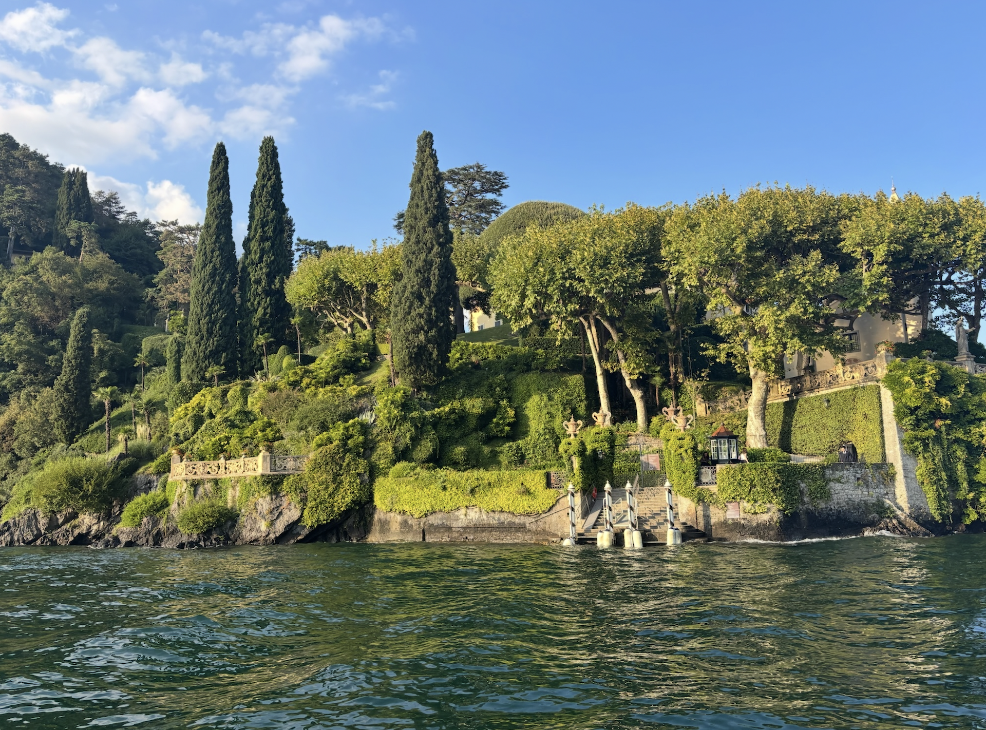 Green hillside with large trees and cypress trees, a white building with a decorative stone wall, and water in the foreground.