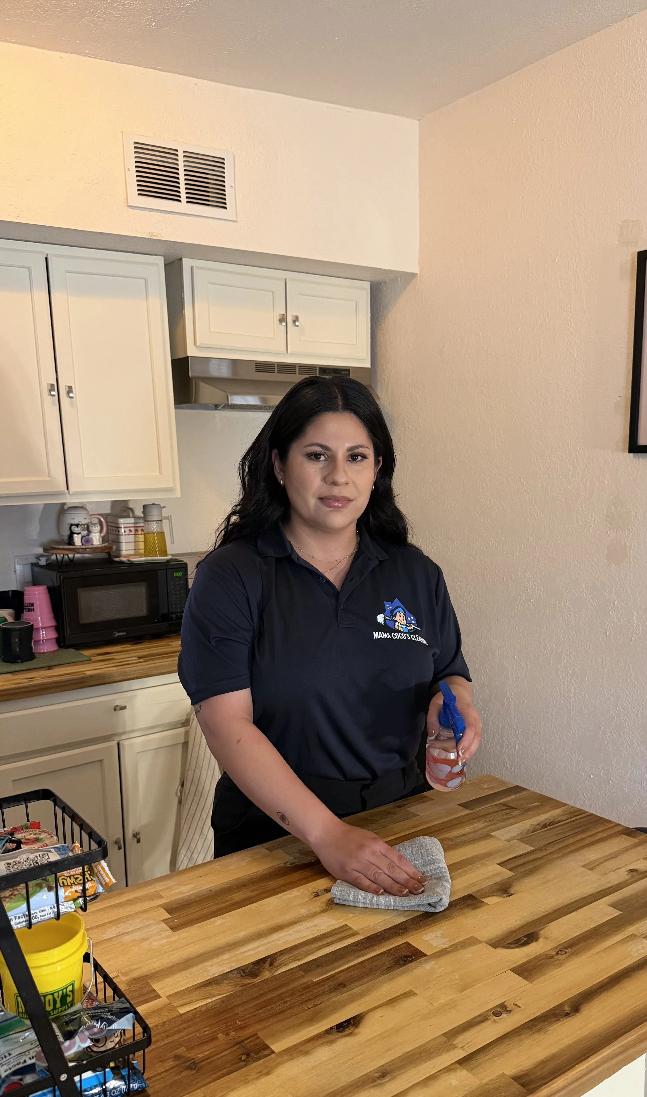 Woman with dark hair wearing a black polo shirt cleaning a wooden kitchen counter with a cloth and holding a spray bottle.