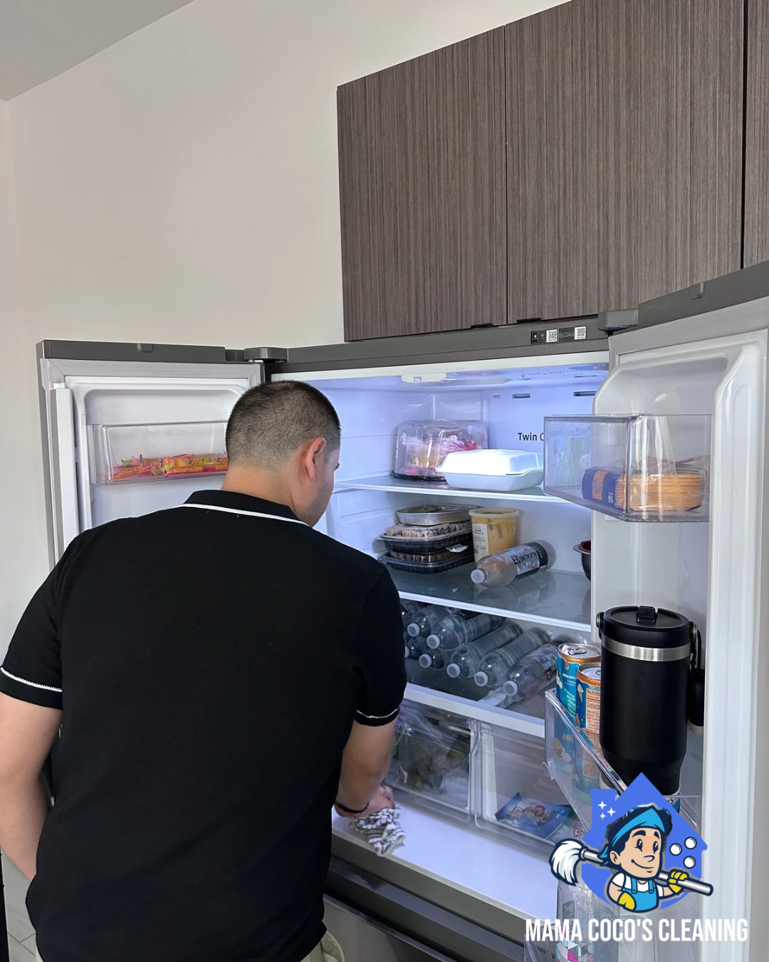 Man cleaning a refrigerator in a kitchen. The open fridge contains various food items, bottles, and containers. A sticker with a cartoon character and the text 'Mama Coco's Cleaning' is on the lower right corner.