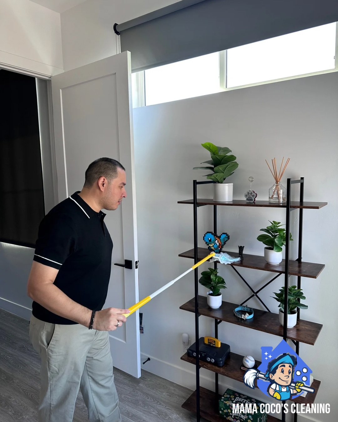 A man is cleaning a houseplant with a long-handled duster while standing next to a wooden bookshelf with potted plants, decorative items, and electronics.