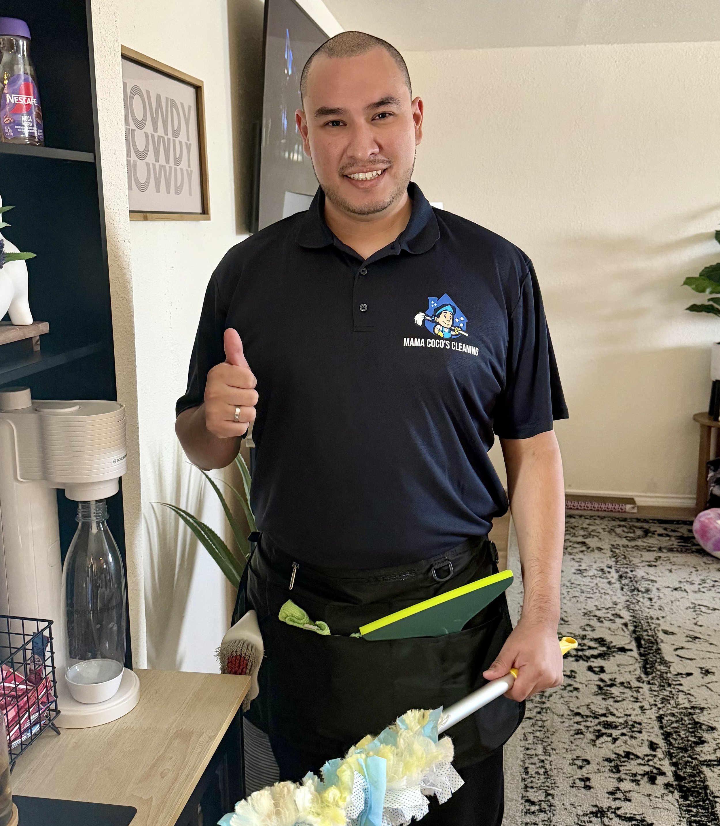 A man with a shaved head and a slight beard smiling and giving a thumbs-up while holding a flower bouquet and cleaning tools. He is wearing a black polo shirt with a logo that reads 'Mama Coco's Cleaning'.