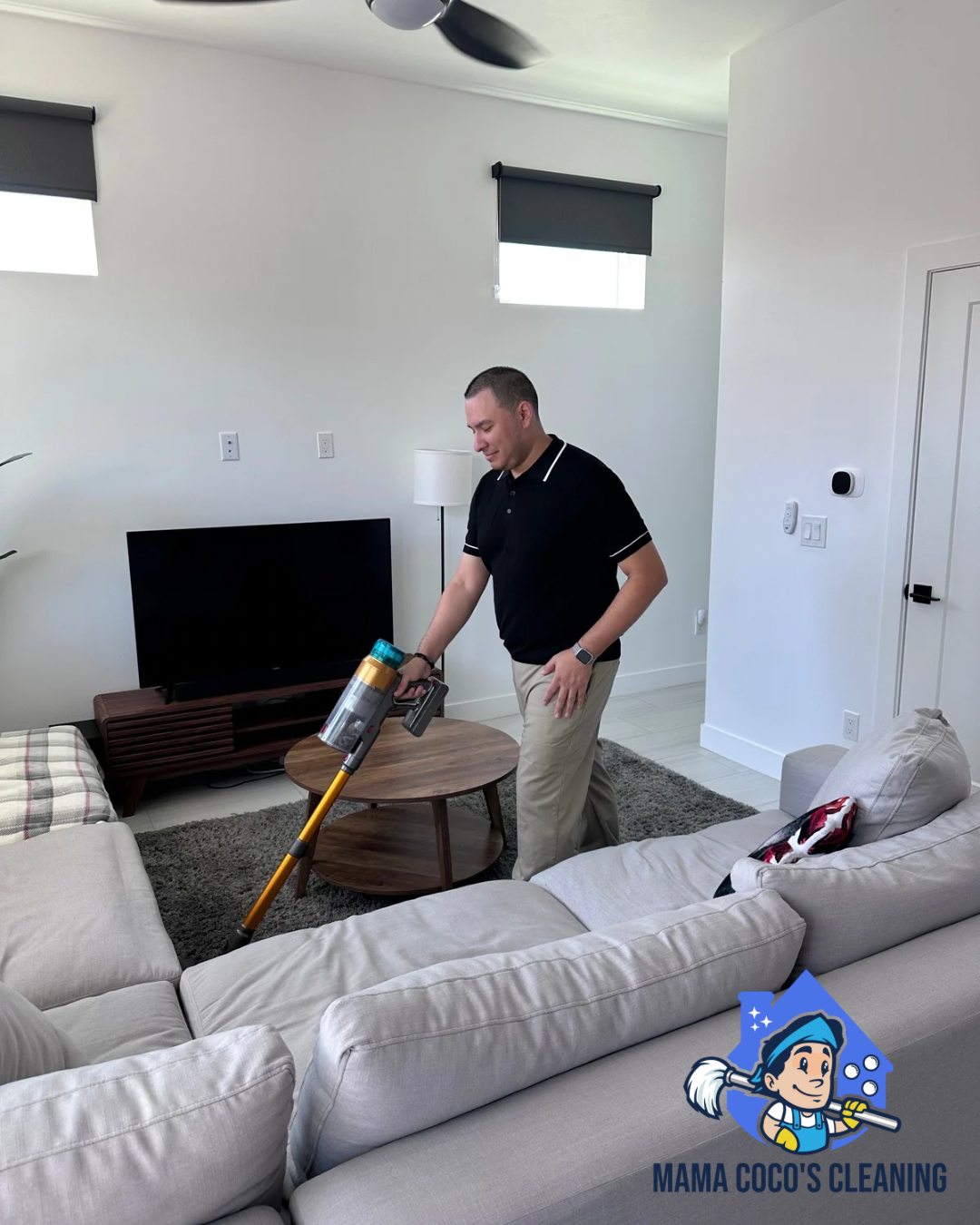 A man vacuuming a living room with beige couches, a coffee table, a TV on a wooden stand, and two windows with gray blinds