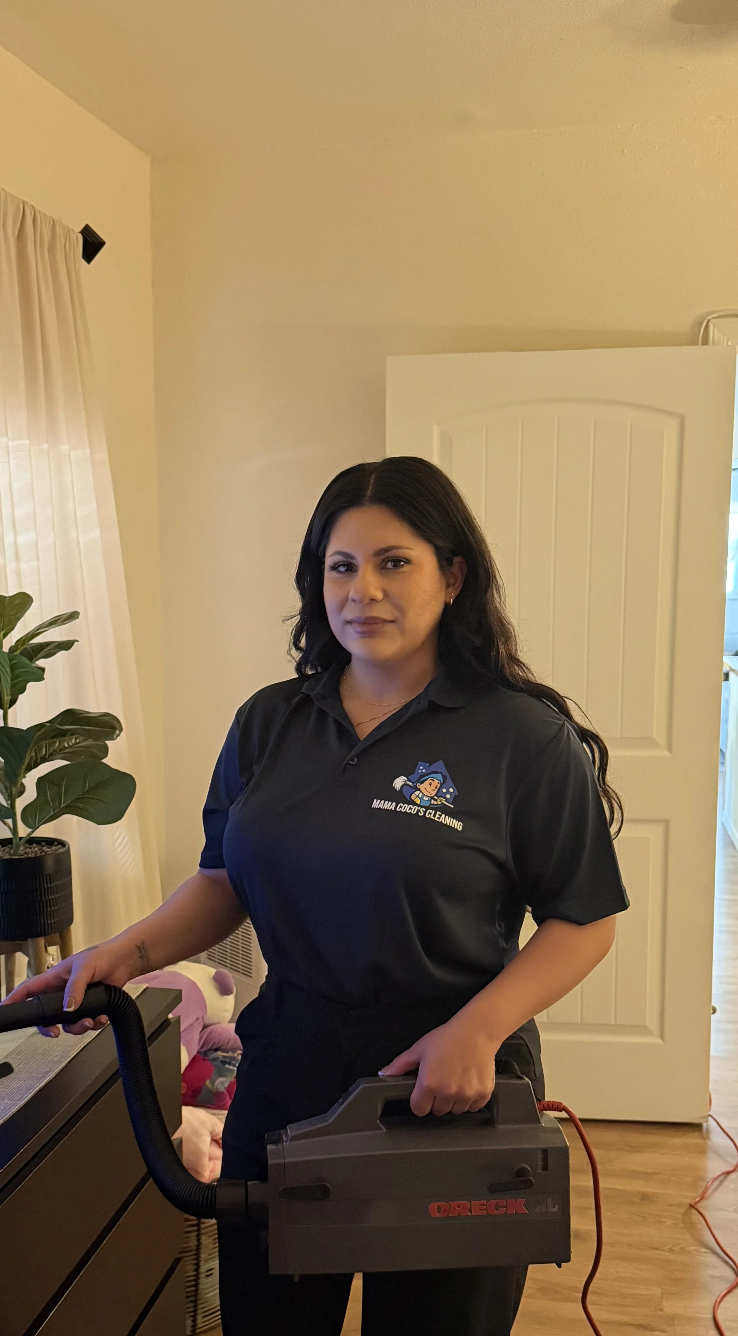 Woman with long dark hair wearing a black shirt with a cleaning logo, holding a black ORECK vacuum cleaner, standing indoors near a door and window with curtains, a plant, and some toys in the background.