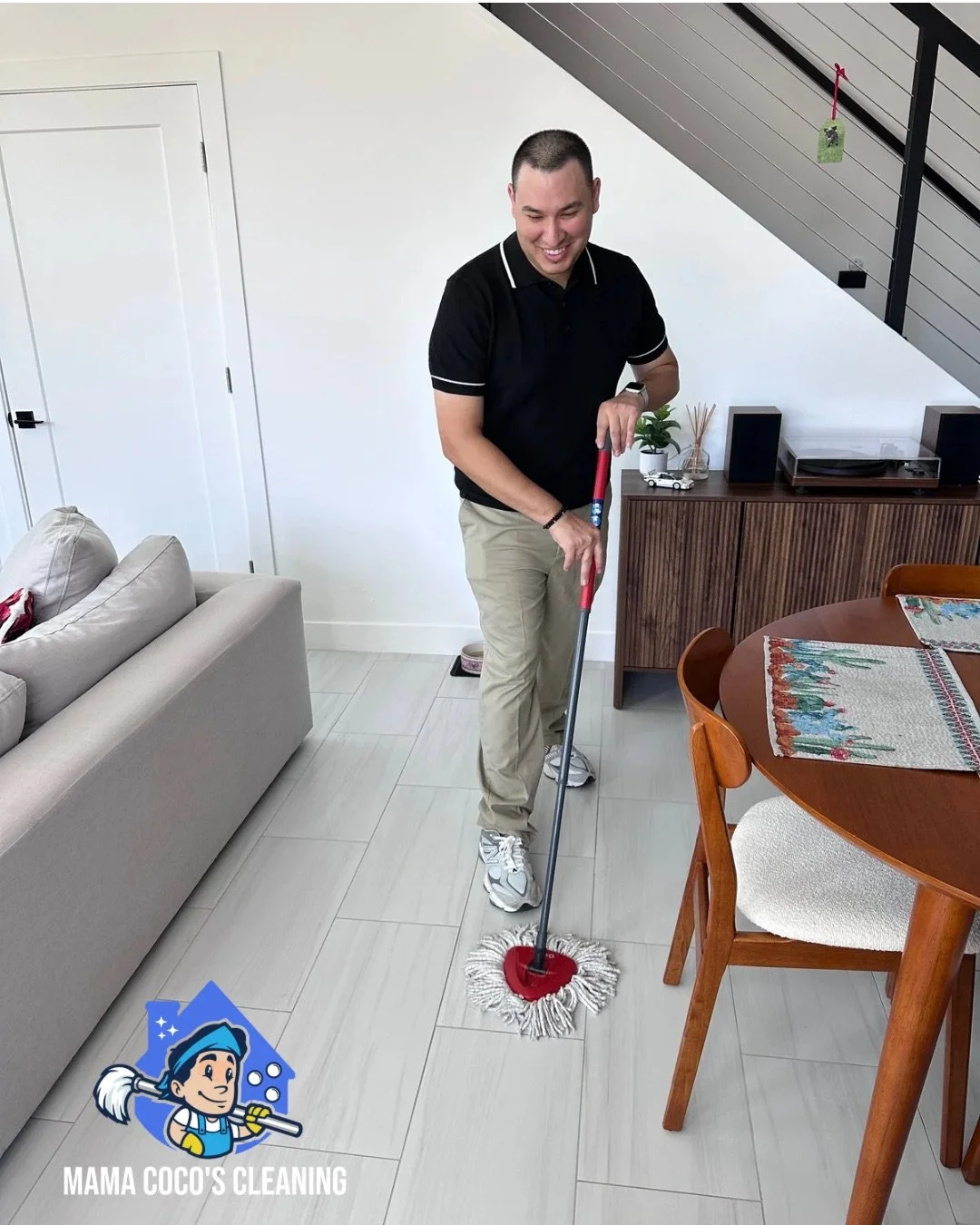 A man cleaning the floor with a mop in a modern living room, smiling, wearing a black polo shirt, beige pants, and sneakers. There is a gray sofa, a wooden dining table with a decorative table runner, a wooden sideboard, houseplants, and a staircase 
