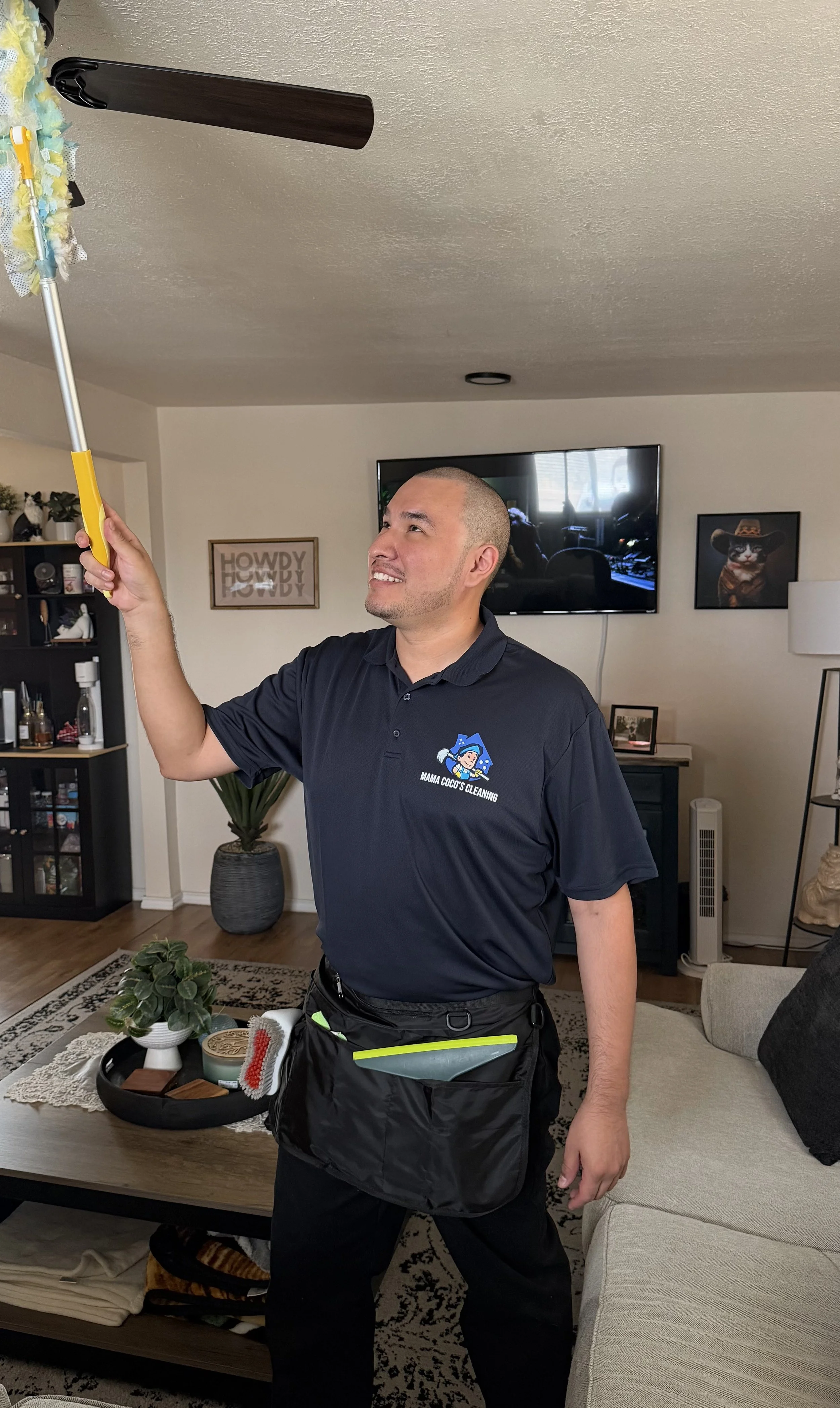 A man with a shaved head smiling in a living room, holding a duster pole with a cleaning cloth attached. He wears a navy blue polo shirt with a logo that says 'Mama Coco's Cleaning' and has a black apron or bag at his waist. The room features a flat-
