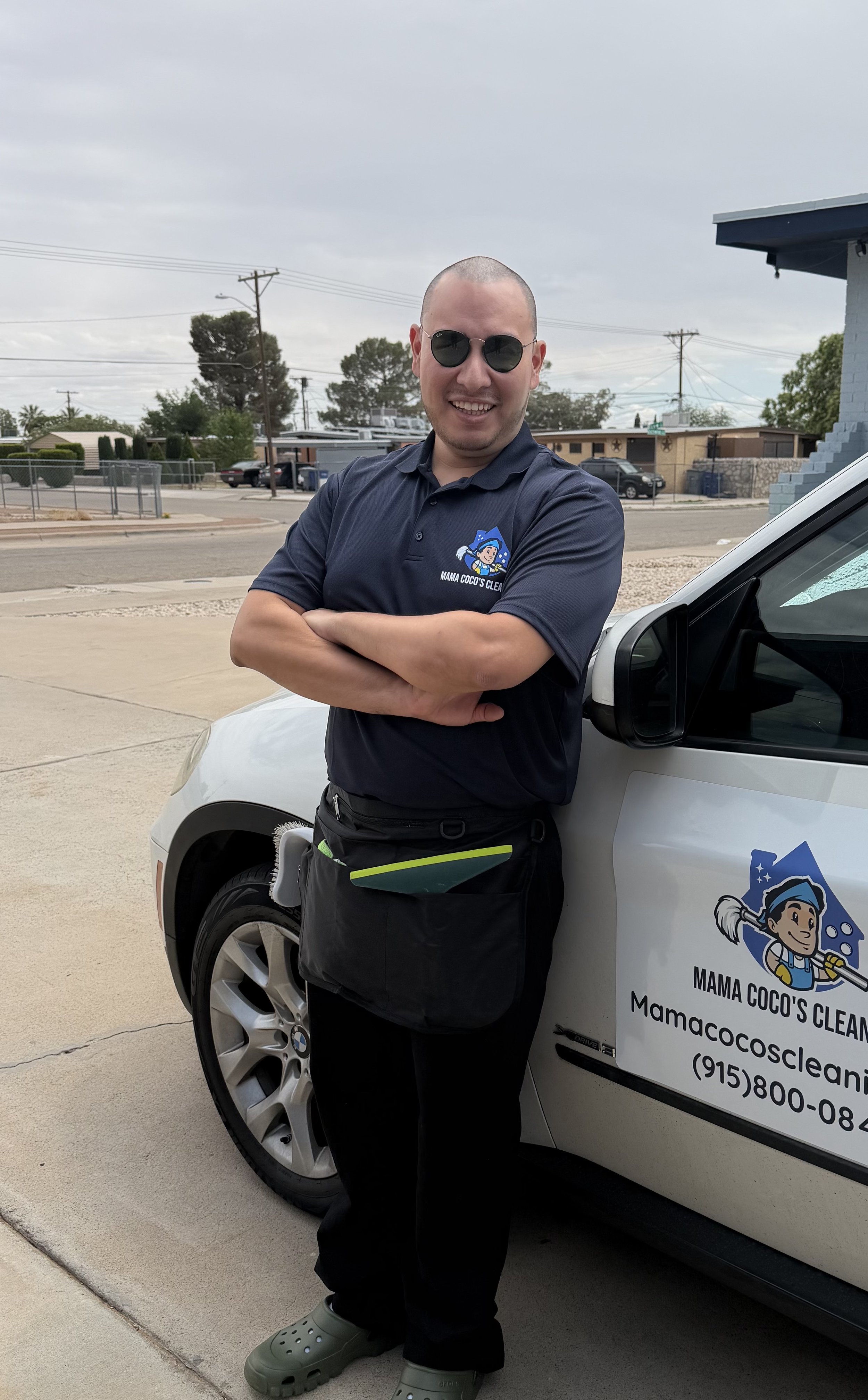 A smiling man in dark sunglasses and a navy polo shirt with a logo stands with arms crossed next to a cleaning service vehicle on a street. The vehicle has the company name 'Mama Coco's Cleaning' and a cartoon logo of a character with a broom.