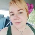 A young woman with blonde hair and a flower in her hair, taking a selfie outdoors on a sunny day.