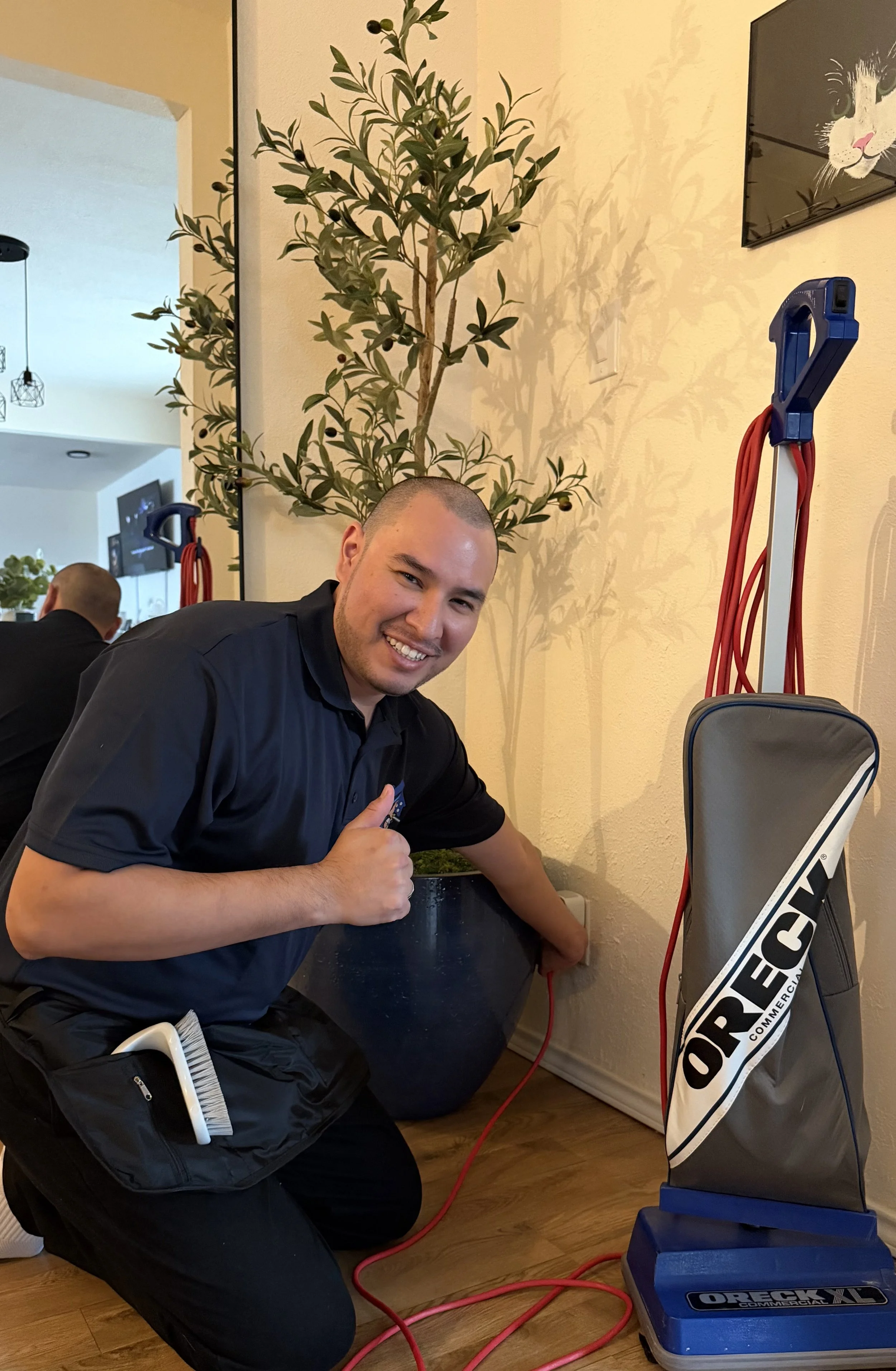 A man with short hair and a goatee smiling while giving a thumbs-up, kneeling next to a treadmill in a room with a light-colored wall, a mirror, and a potted plant, with exercise equipment and black and red cords nearby.