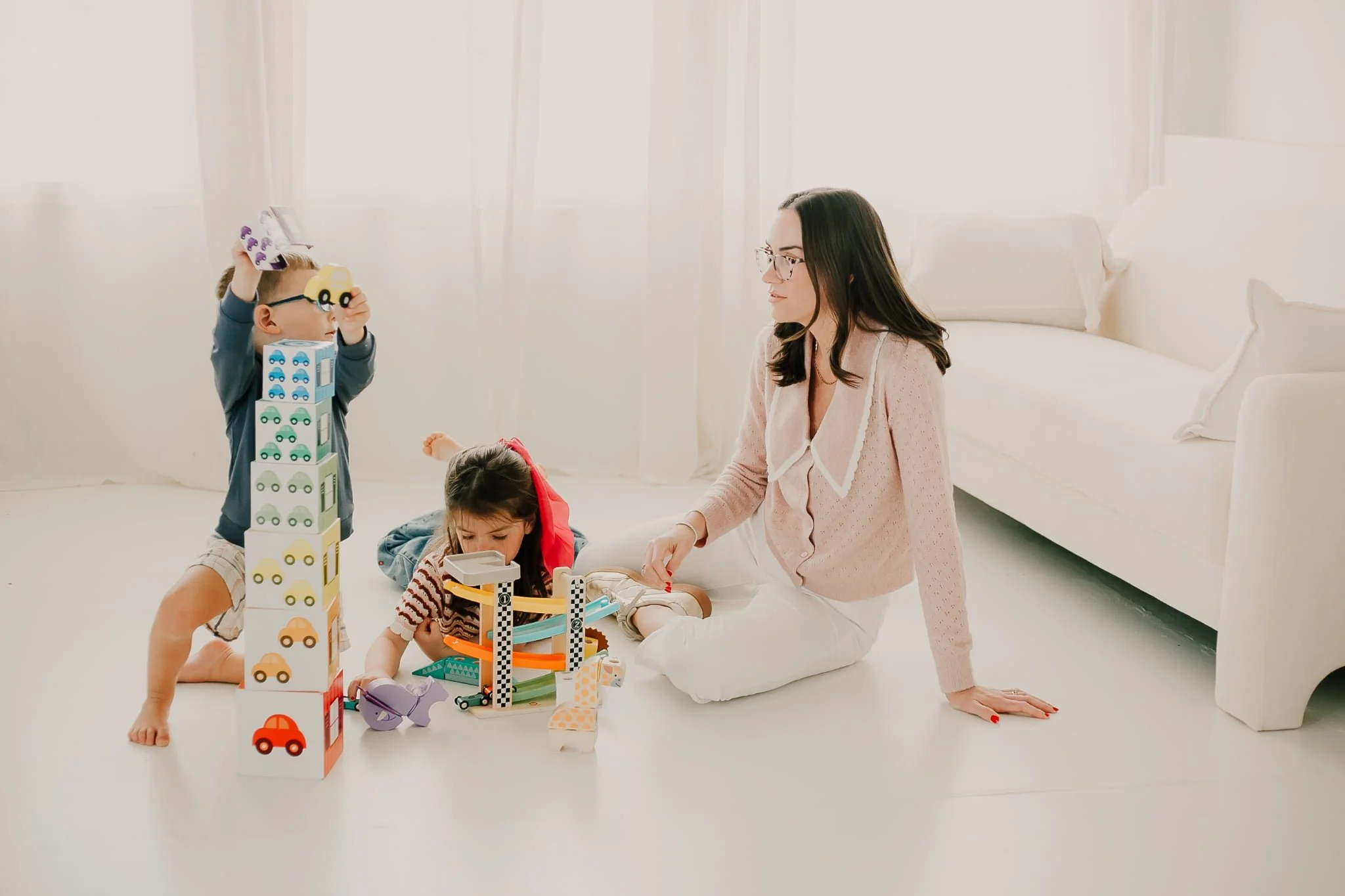 A woman with dark hair, glasses, and a pink cardigan sits on the floor playing with two children; one boy with glasses standing and stacking toy blocks, and one girl with dark hair sitting and playing with toys in front of her, in a bright, minimalistic room with a white sofa, white walls, and sheer white curtains.