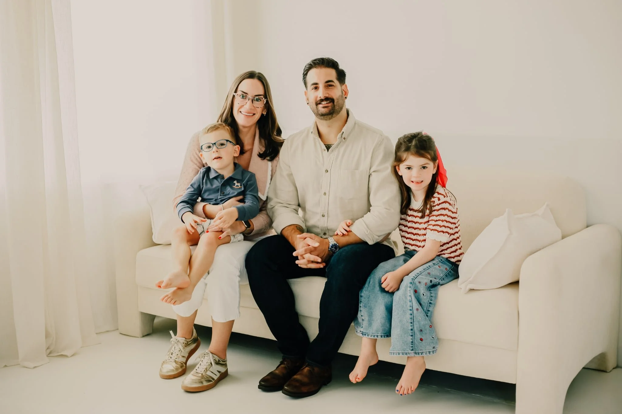 A family of five sitting on a white sofa in a bright room, smiling at the camera. The mother, with glasses, is holding a young boy with glasses. A man with a beard is sitting next to a girl with long hair and a red ribbon.