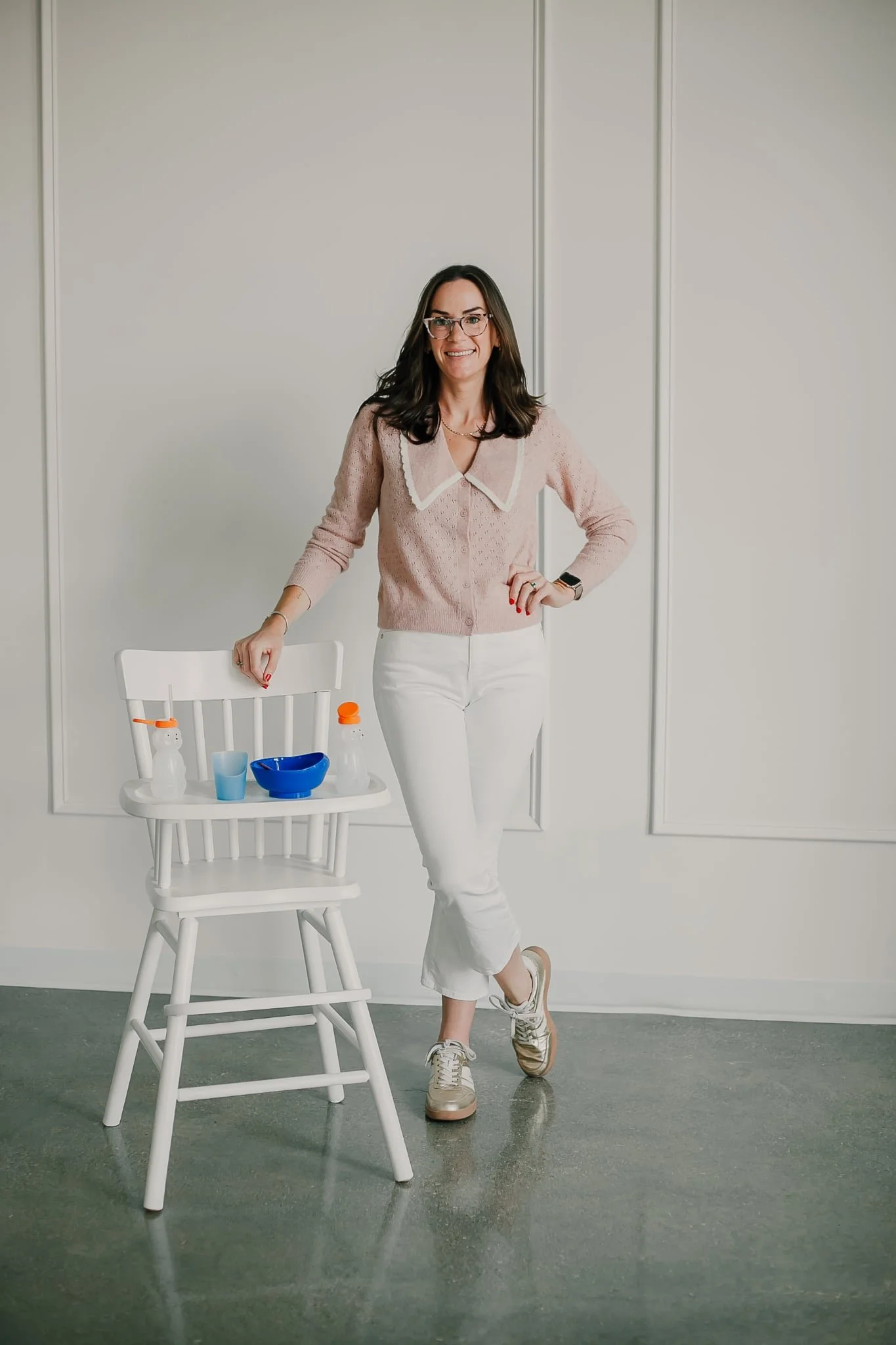 A woman with dark hair, glasses, and light skin, wearing a pink sweater with a white collar and white pants, standing in front of a plain white wall, smiling and posing with one hand on her hip. There is a white chair with colorful bottles and a blue bowl on it in front of her, and the floor is made of gray concrete.
