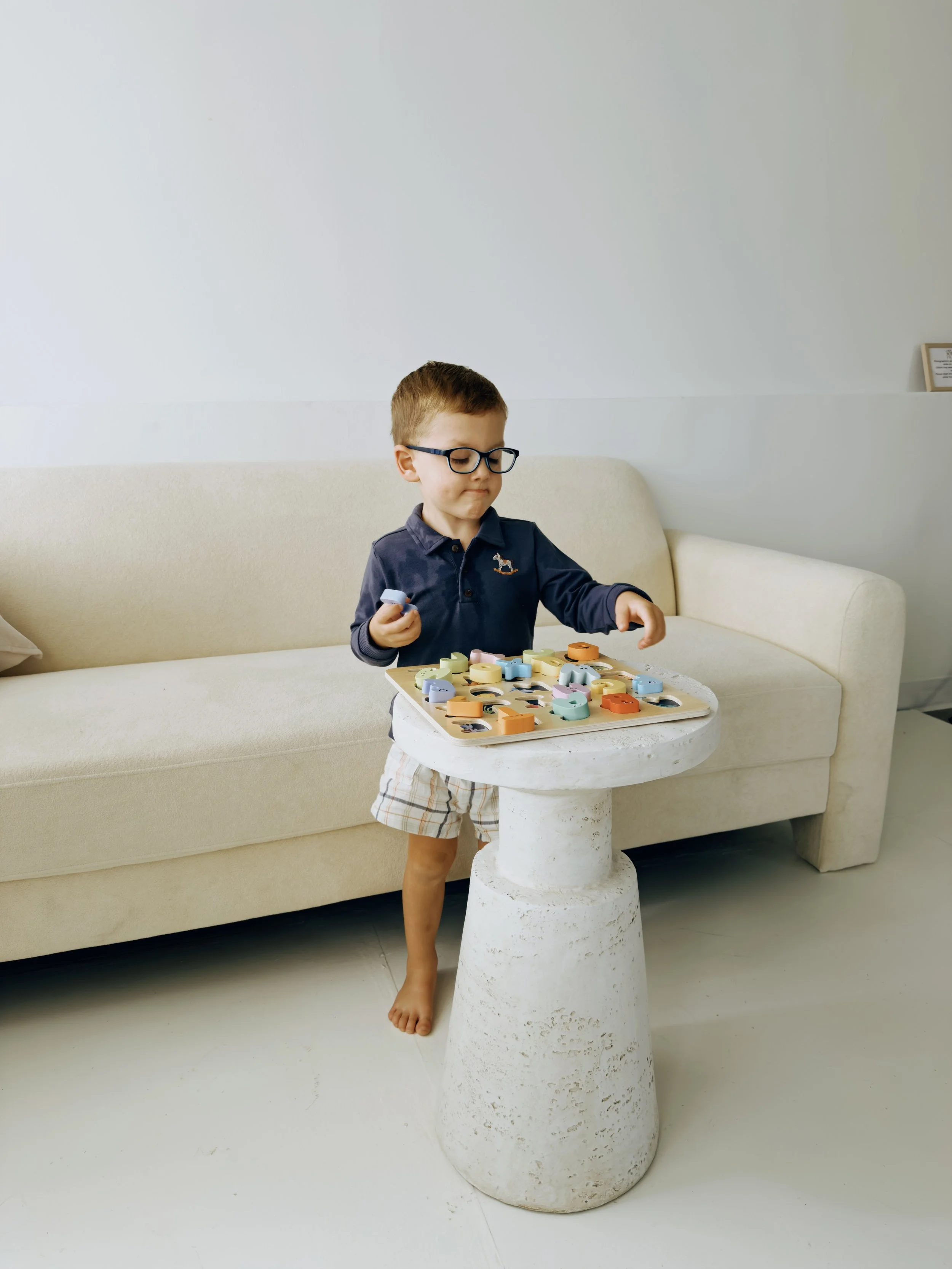 A young boy with glasses is playing with colorful wooden alphabet letters on a small two-legged white table in a living room. He is standing barefoot, wearing a navy blue shirt with a small design on the chest and plaid shorts. Behind him is a beige curved sofa against a light-colored wall.