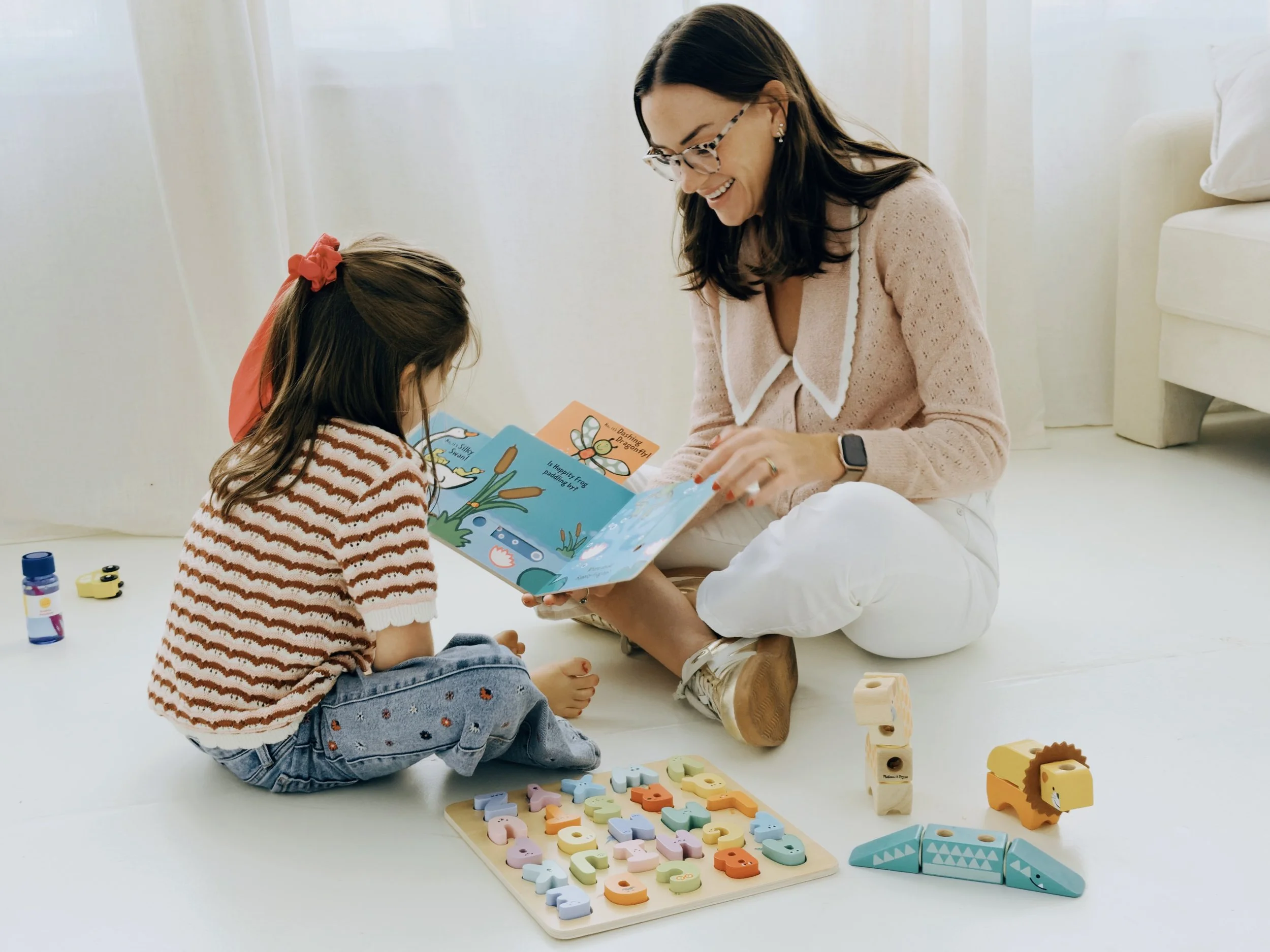 A woman and a girl sitting on the floor playing with colorful wooden alphabet puzzles and toys in a bright room with white curtains.